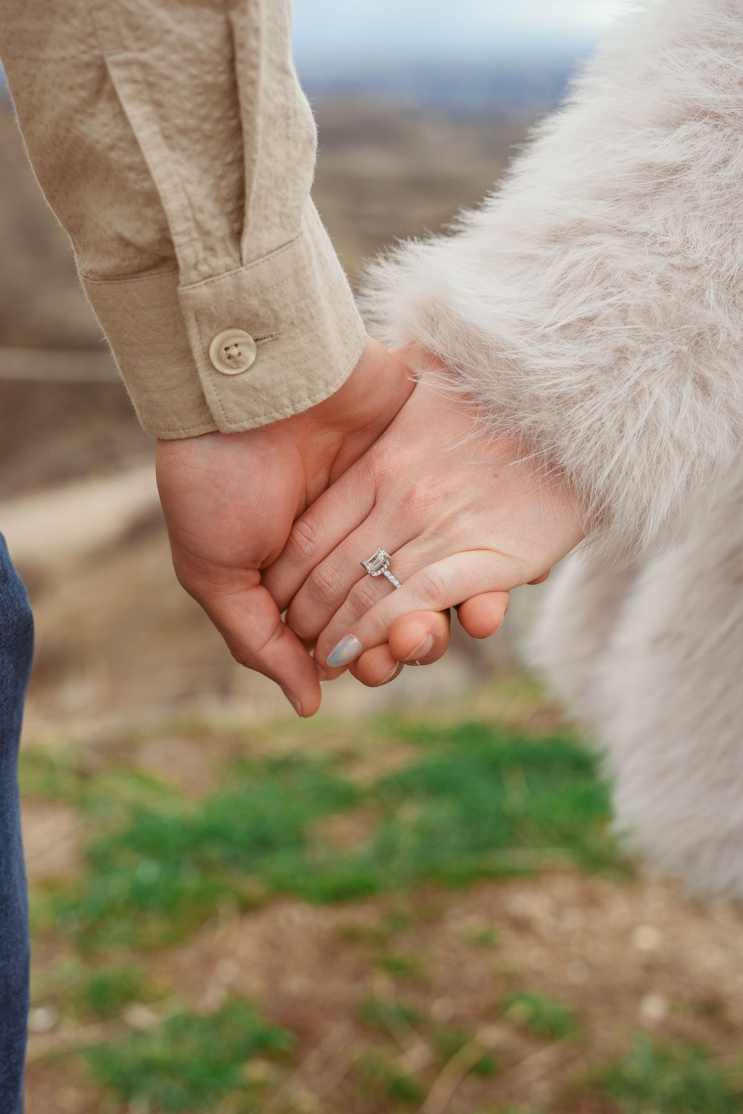Close up of hands holding together showing engagement ring against white fur background.