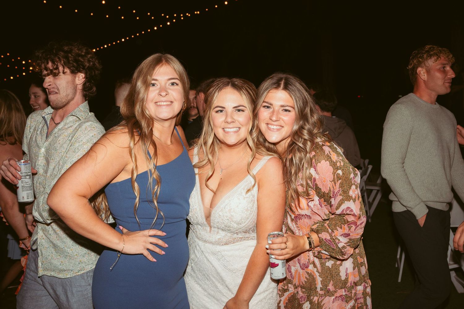 Three friends in formal attire pose together at an outdoor evening event under string lights.