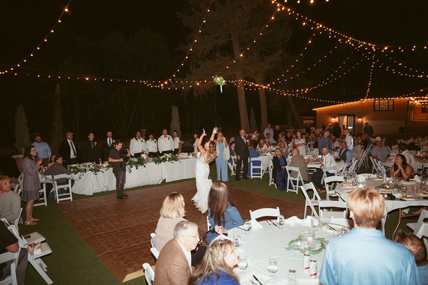 Wedding reception under twinkling lights with guests seated at tables watching a couple dance on a patio.