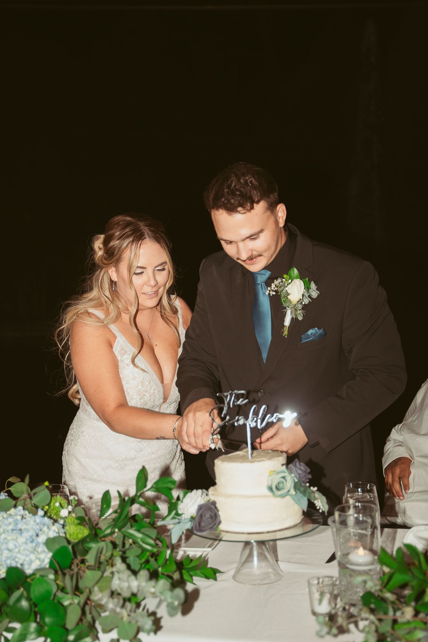 Couple cutting their white wedding cake together with greenery decorations on the table.