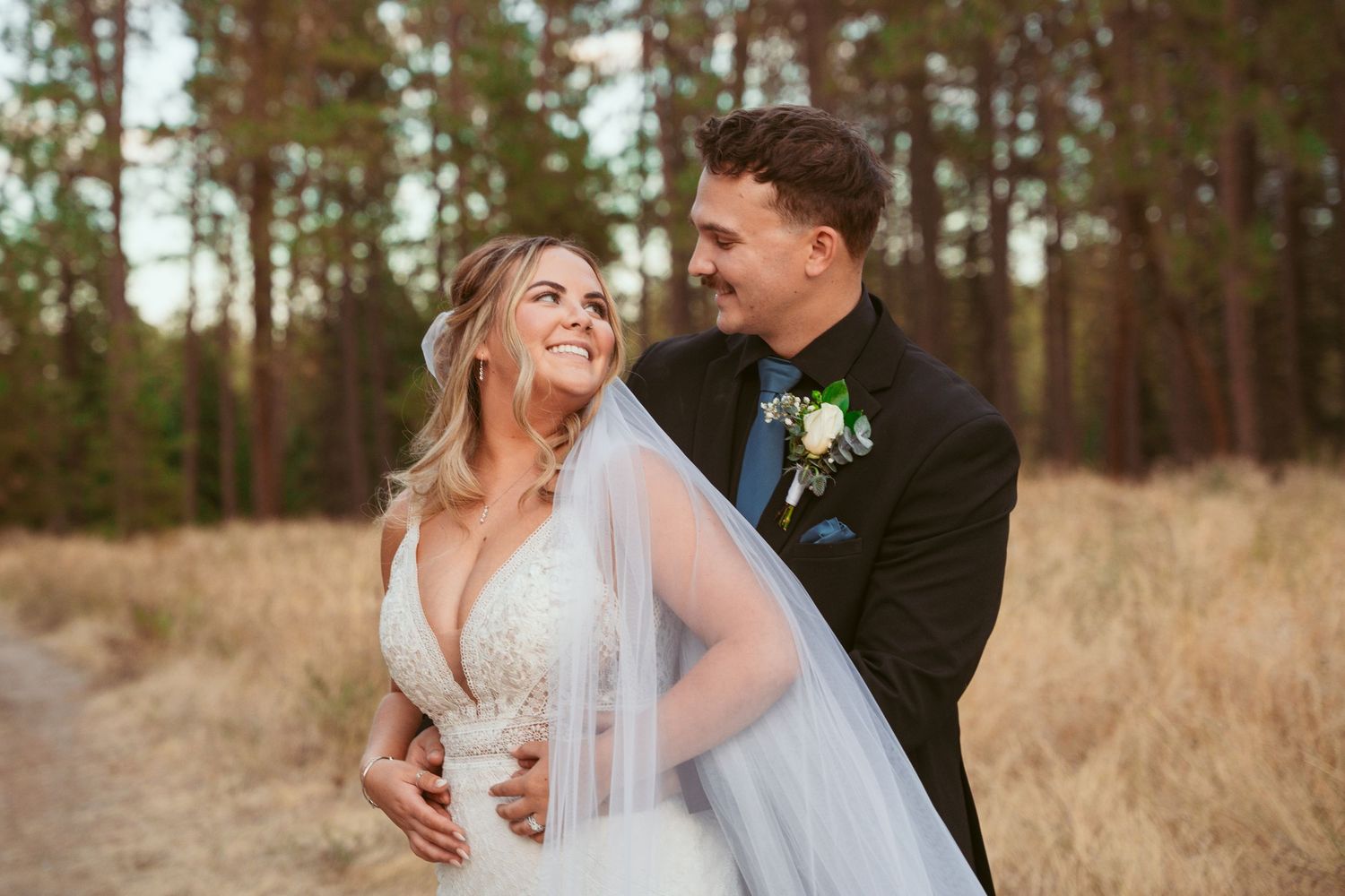 Newlyweds share intimate moment in rustic outdoor setting with pine trees and golden grass.