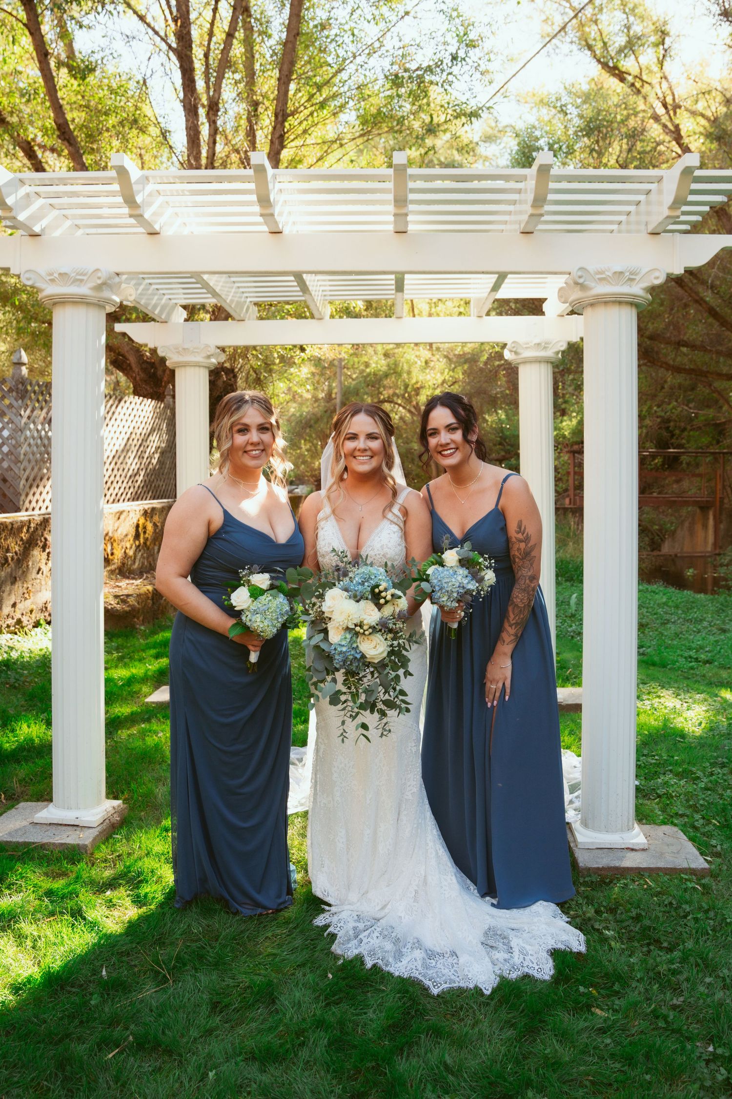 Wedding party in navy blue dresses poses with bride under a white pergola in a garden setting.