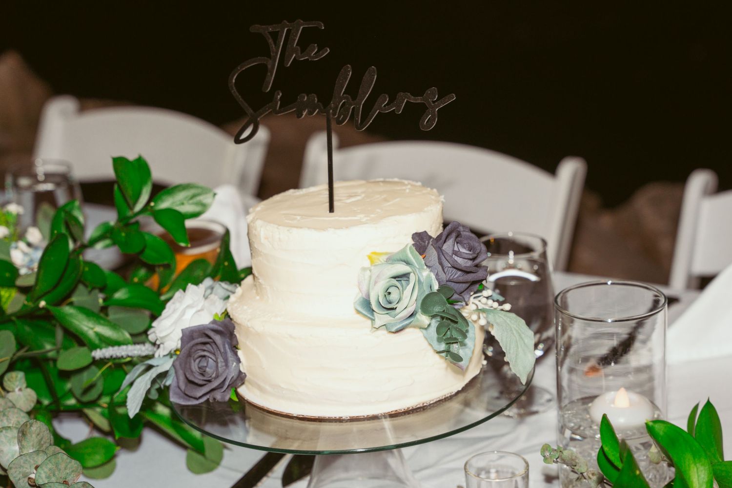 White wedding cake with delicate floral decorations and green foliage sits elegantly on a rustic wooden table.