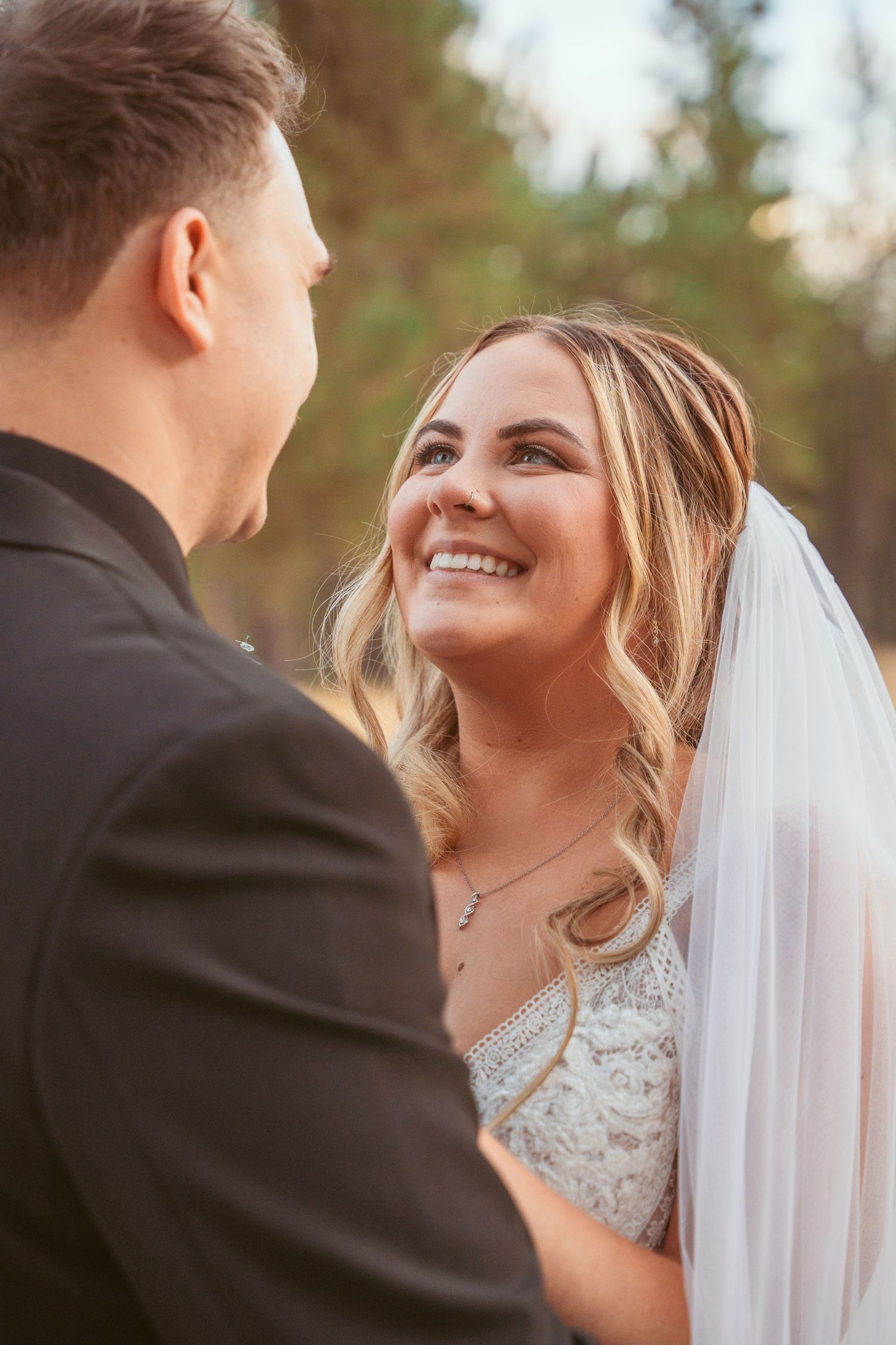 A romantic wedding moment captured between a bride in white lace and groom in a dark suit against a natural outdoor backdrop.