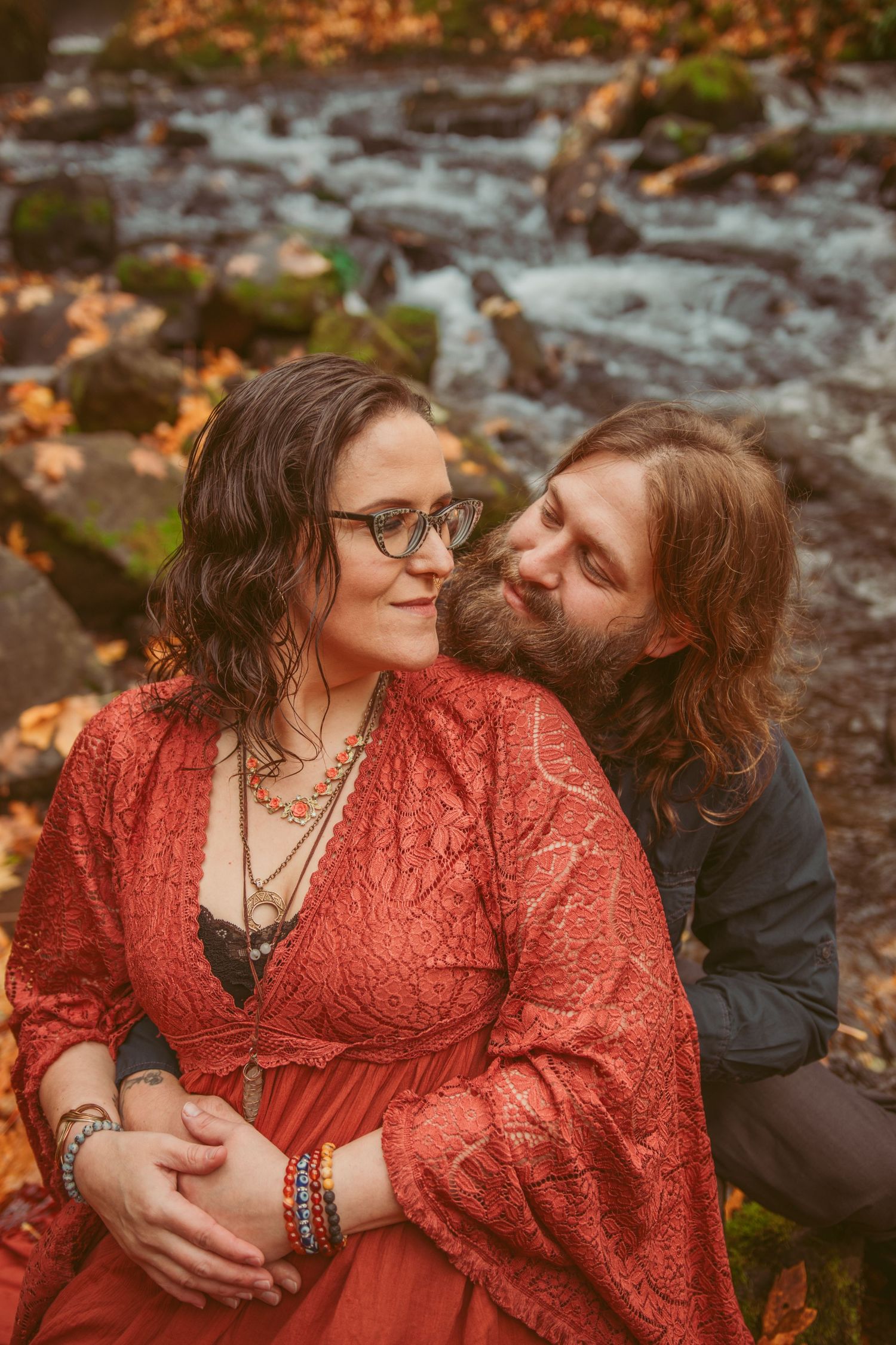 A couple shares a romantic moment by a stream with autumn foliage in the background.