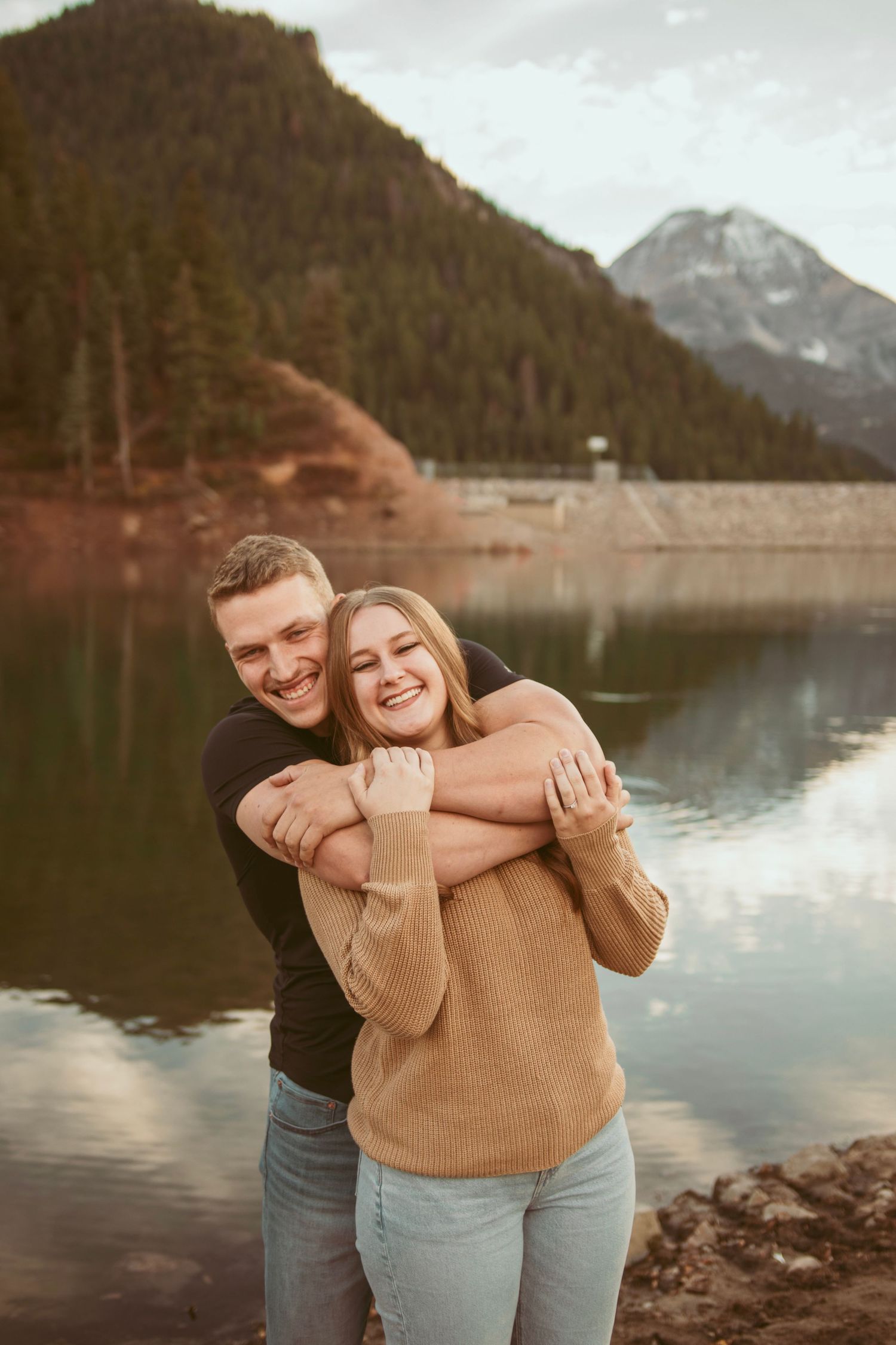 A couple shares playful moments by a mountain lake with evergreen forests in the background.
