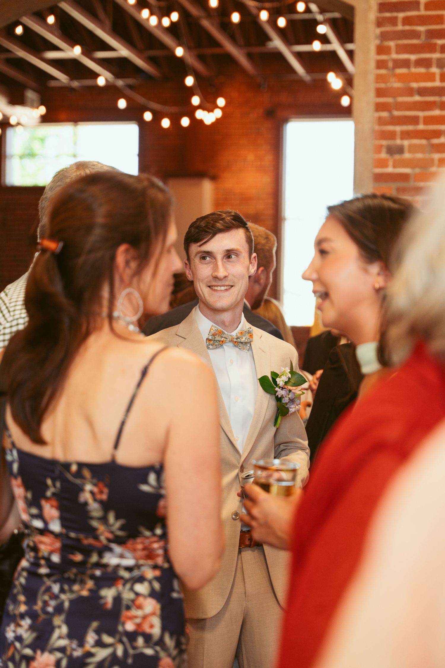 Wedding guests socialize in an industrial-style venue with exposed brick walls and string lights.