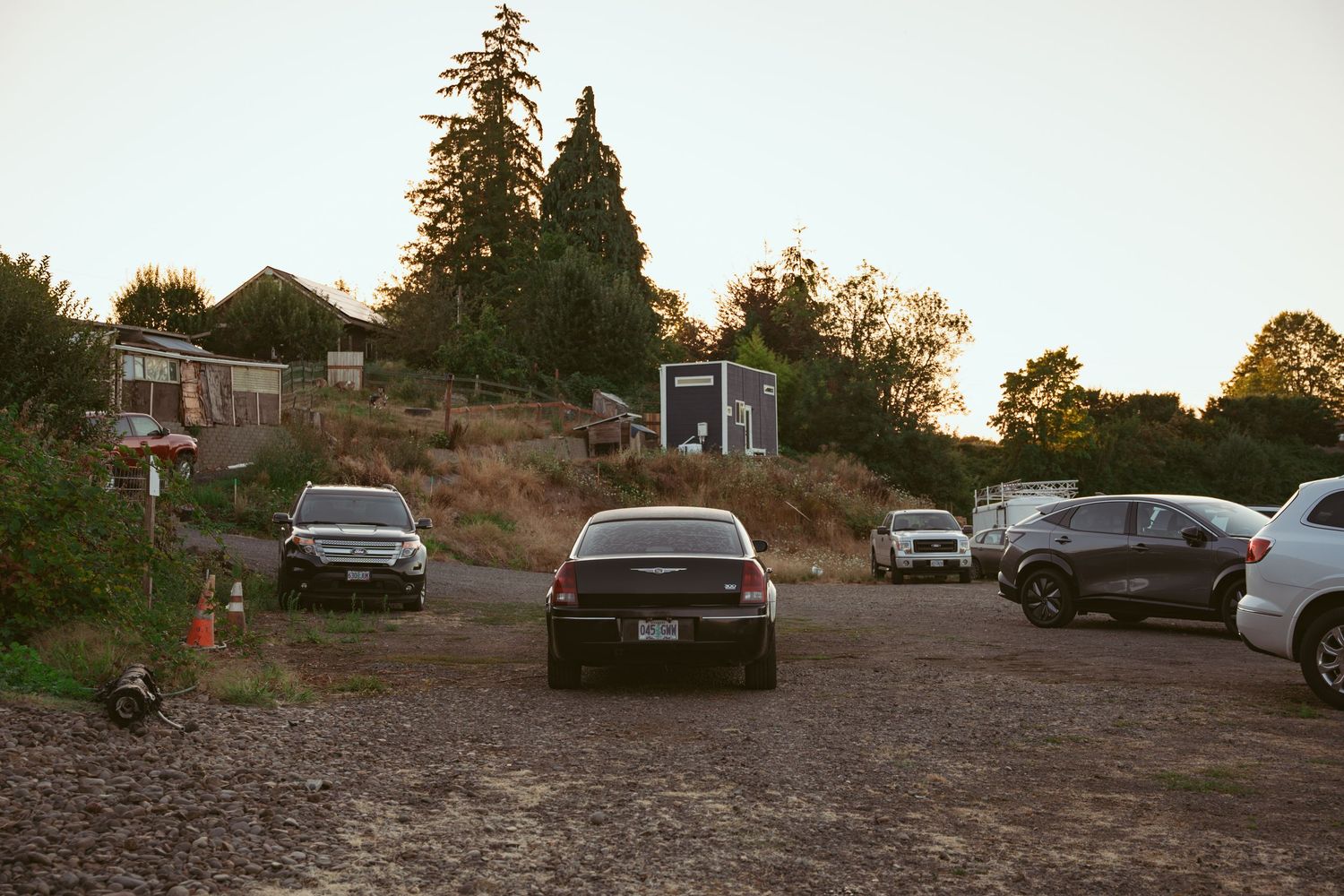 Cars parked on a dirt lot overlooking trees and buildings at sunset.