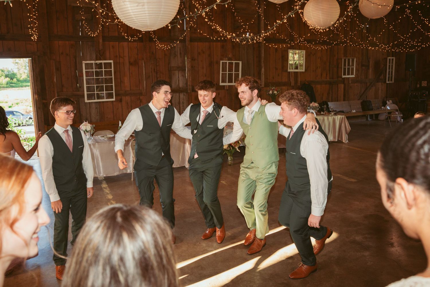 People dancing in formation at a barn reception with twinkling lights overhead.