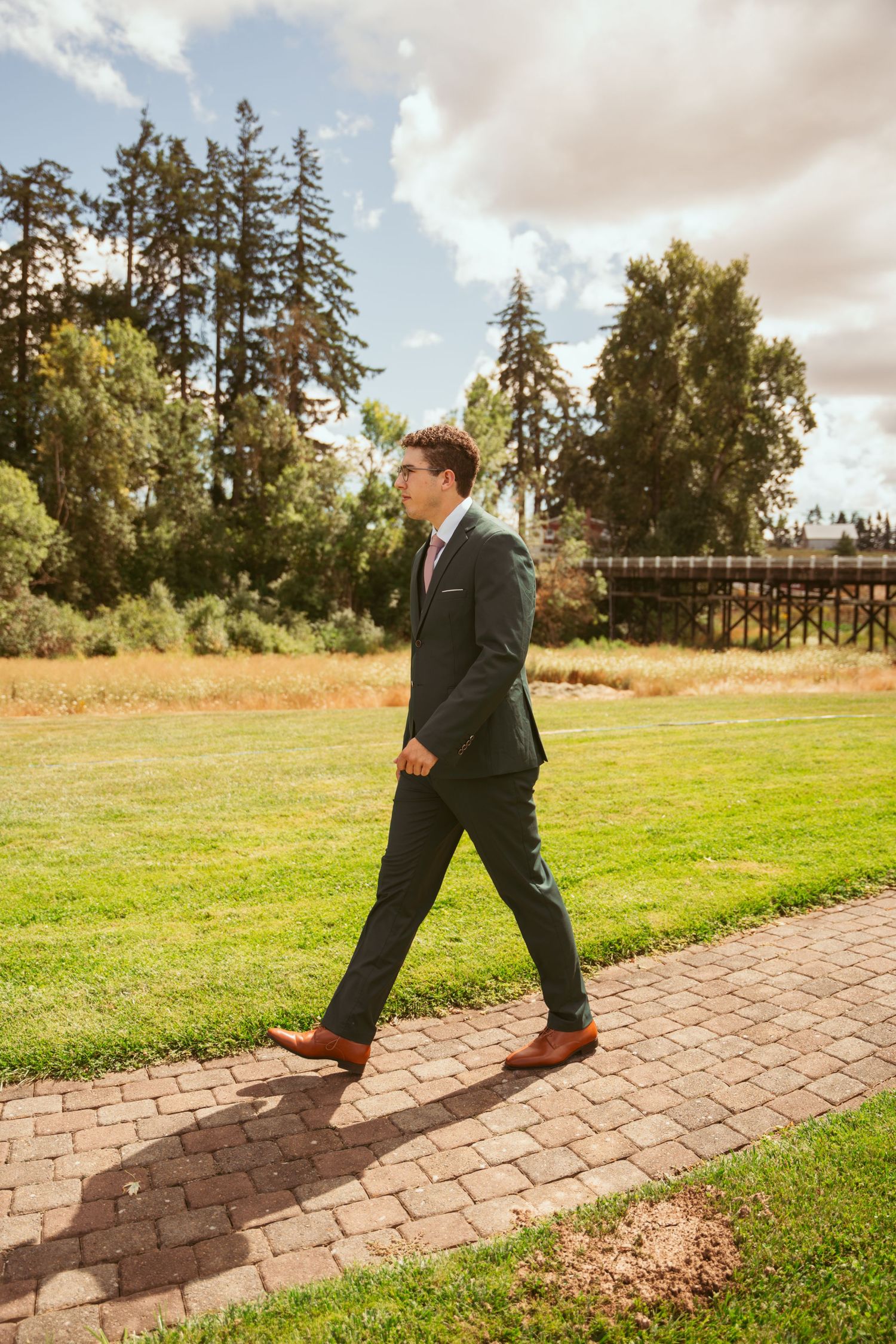Person in dark suit strolls down brick pathway against backdrop of evergreen trees.