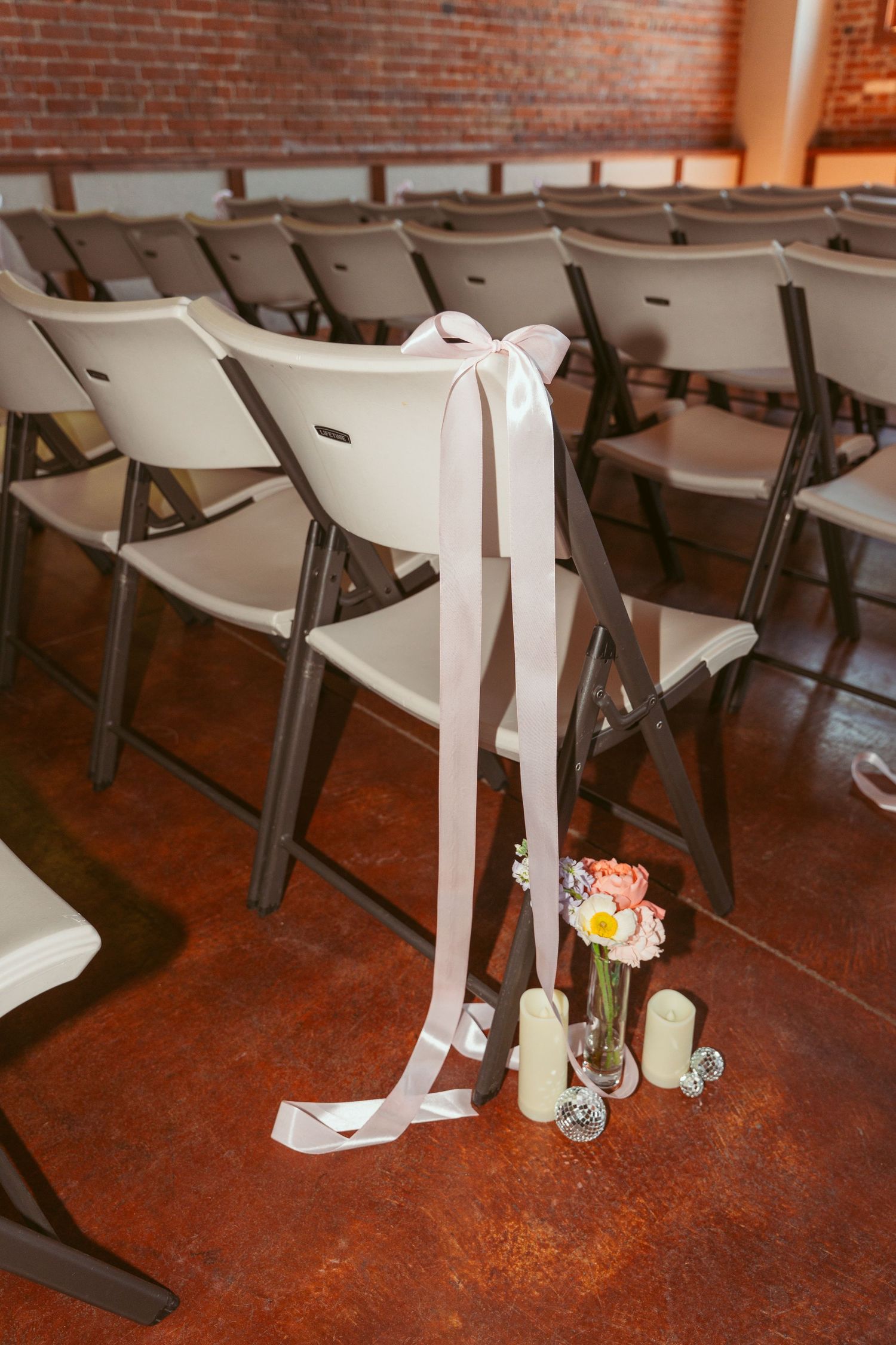 Rows of white folding chairs with ribbon decorations and floral arrangements in a brick-walled venue.