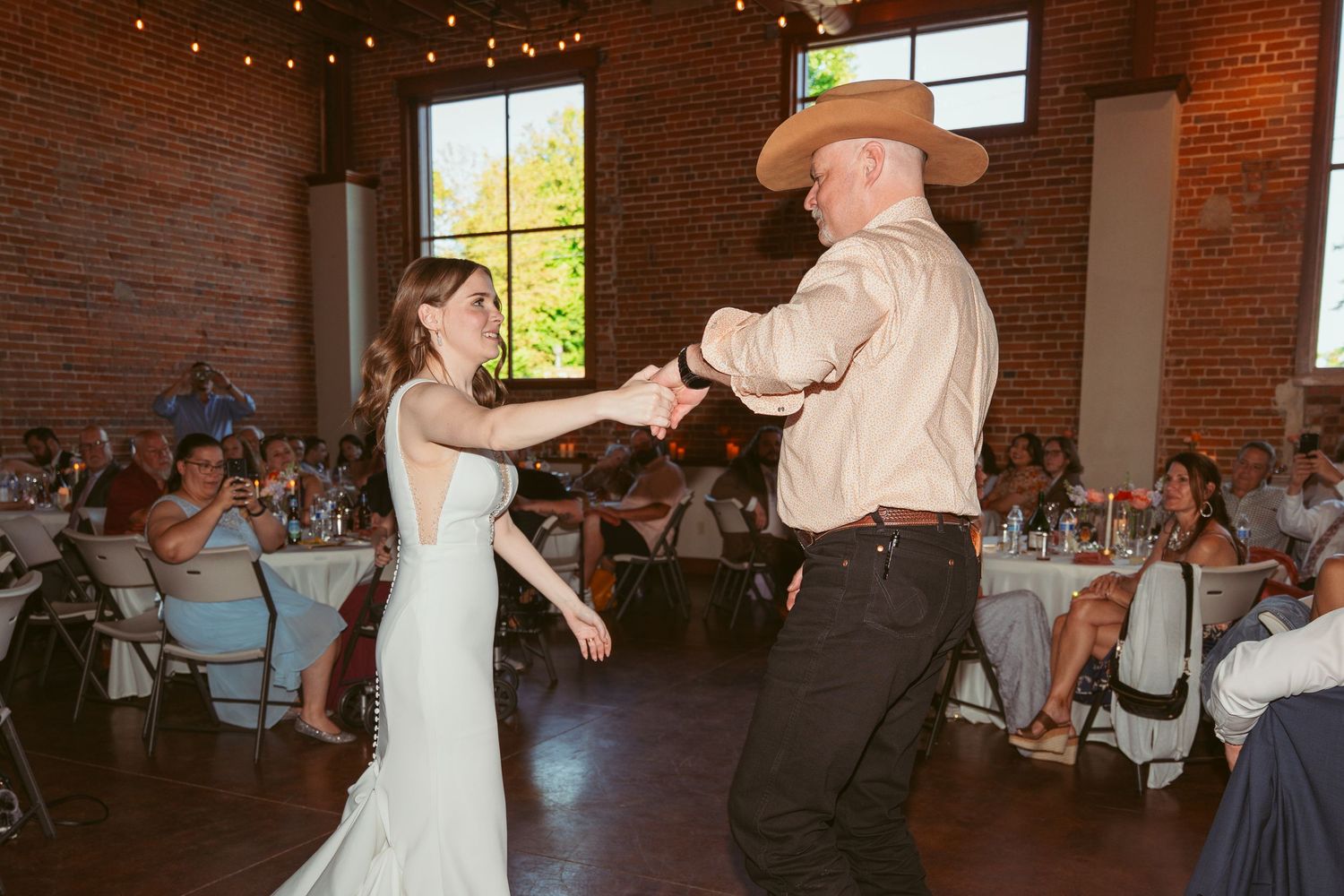 Wedding reception in a rustic brick venue with string lights overhead and guests dancing.