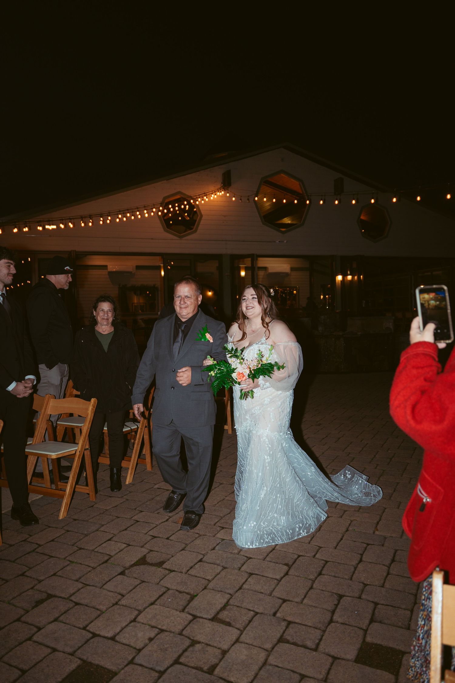 Wedding party members dance on a stone patio under twinkling lights at night.