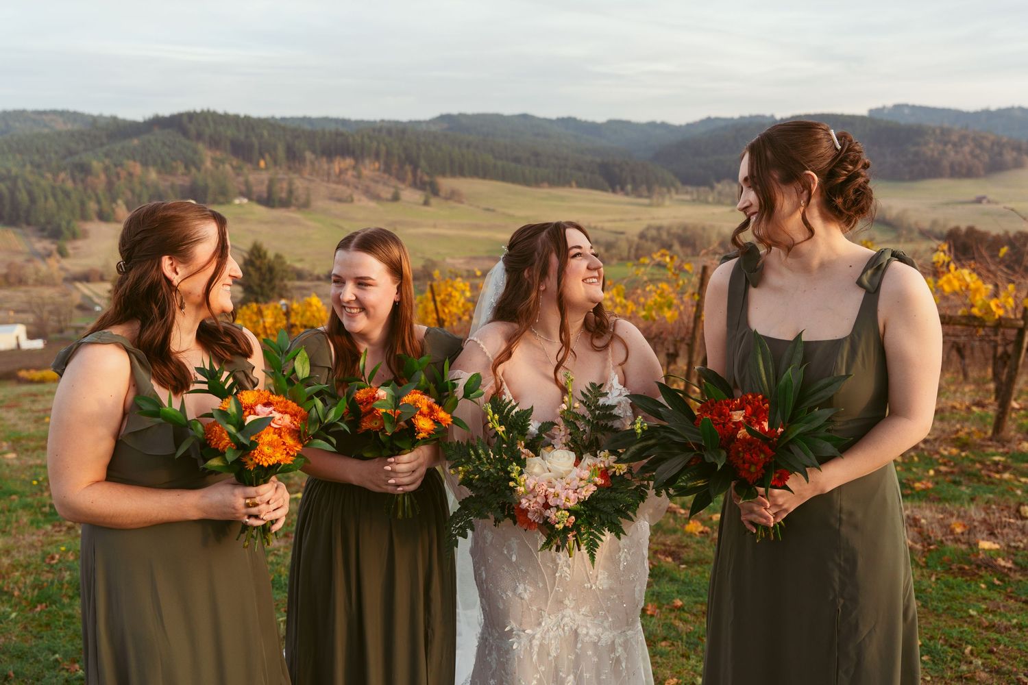 Bridal party in olive green dresses holds autumn-colored bouquets against vineyard backdrop.