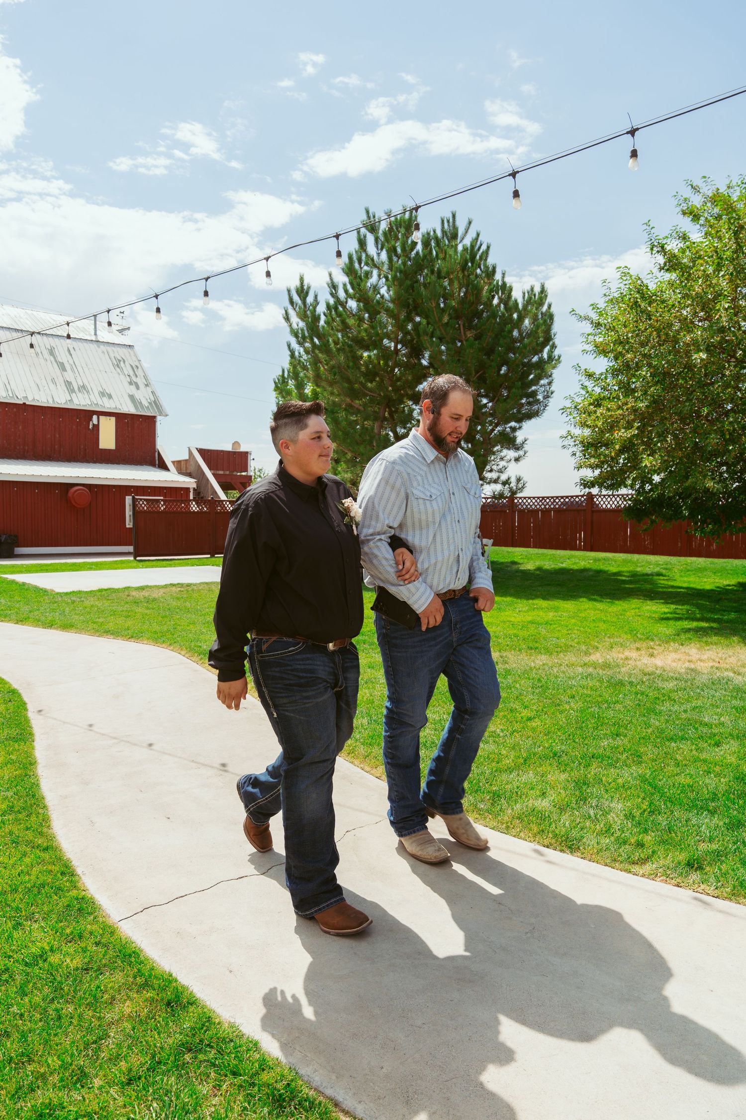 Two people walk along a concrete path near a red barn under string lights on a sunny day.