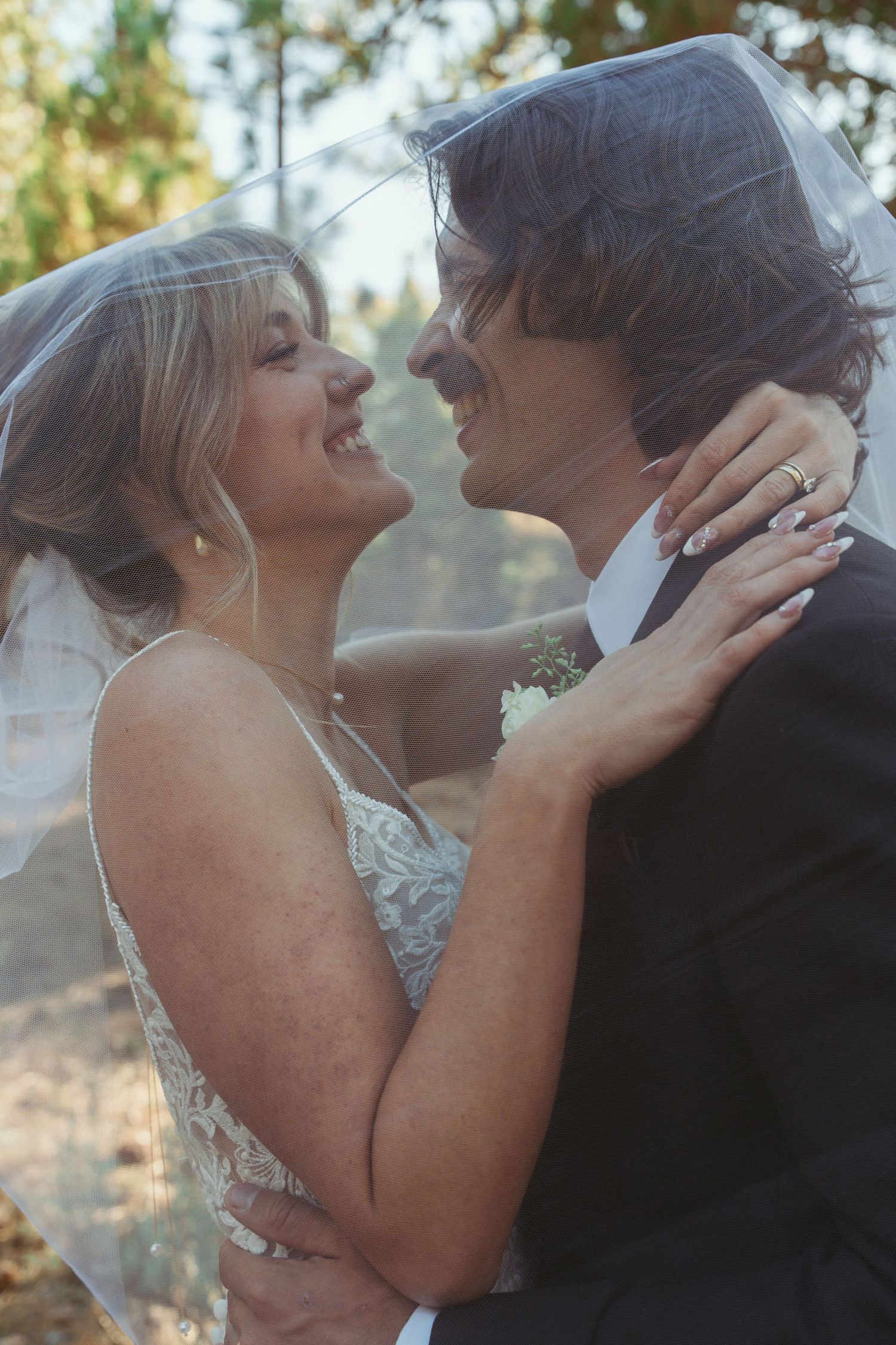 Bride and groom smiling at each other under a wedding veil, embracing outdoors in a romantic forest setting.