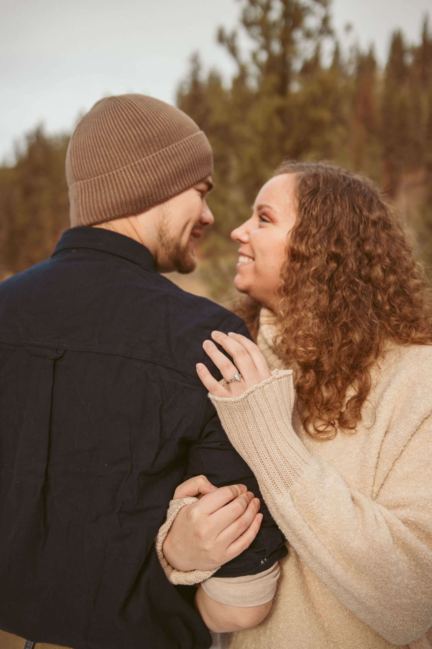 Couple in winter clothing embracing outdoors against a forest backdrop.