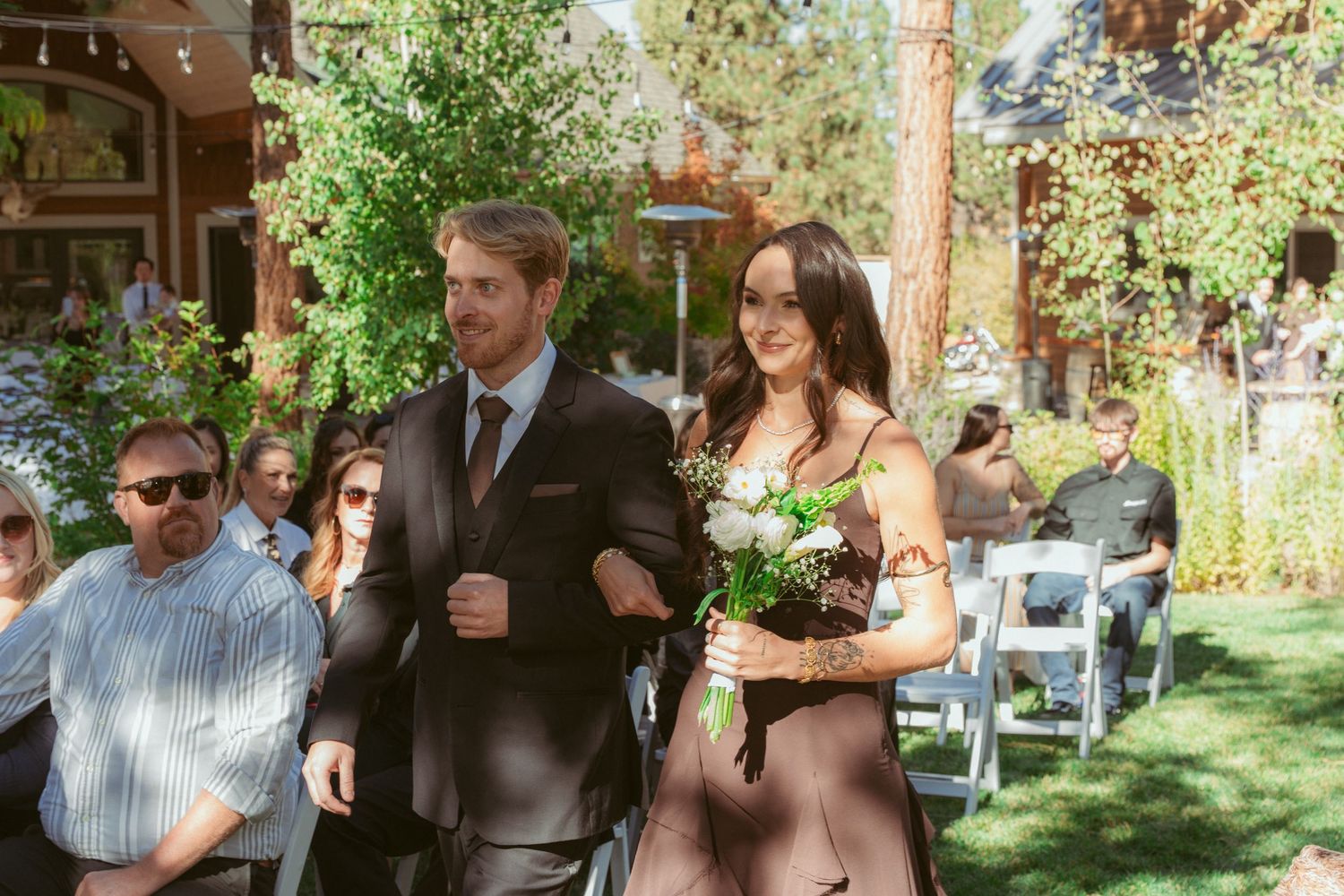 Outdoor wedding ceremony with guests seated in white chairs under trees on a sunny day.