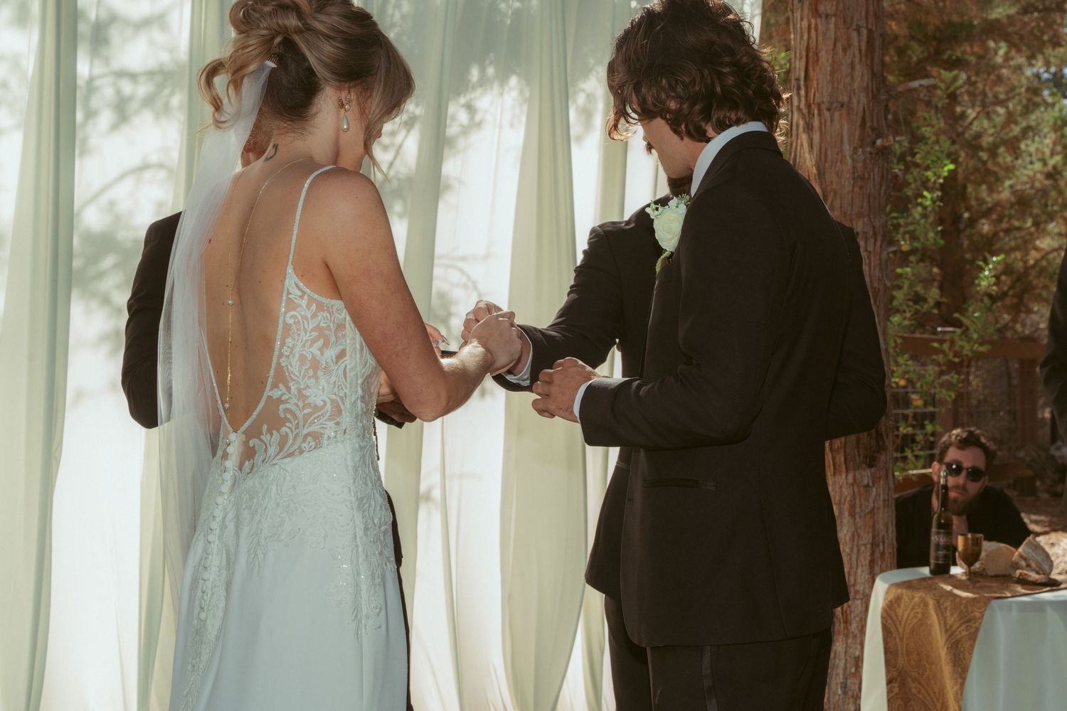 A couple exchanges rings during their wedding ceremony beneath towering trees.