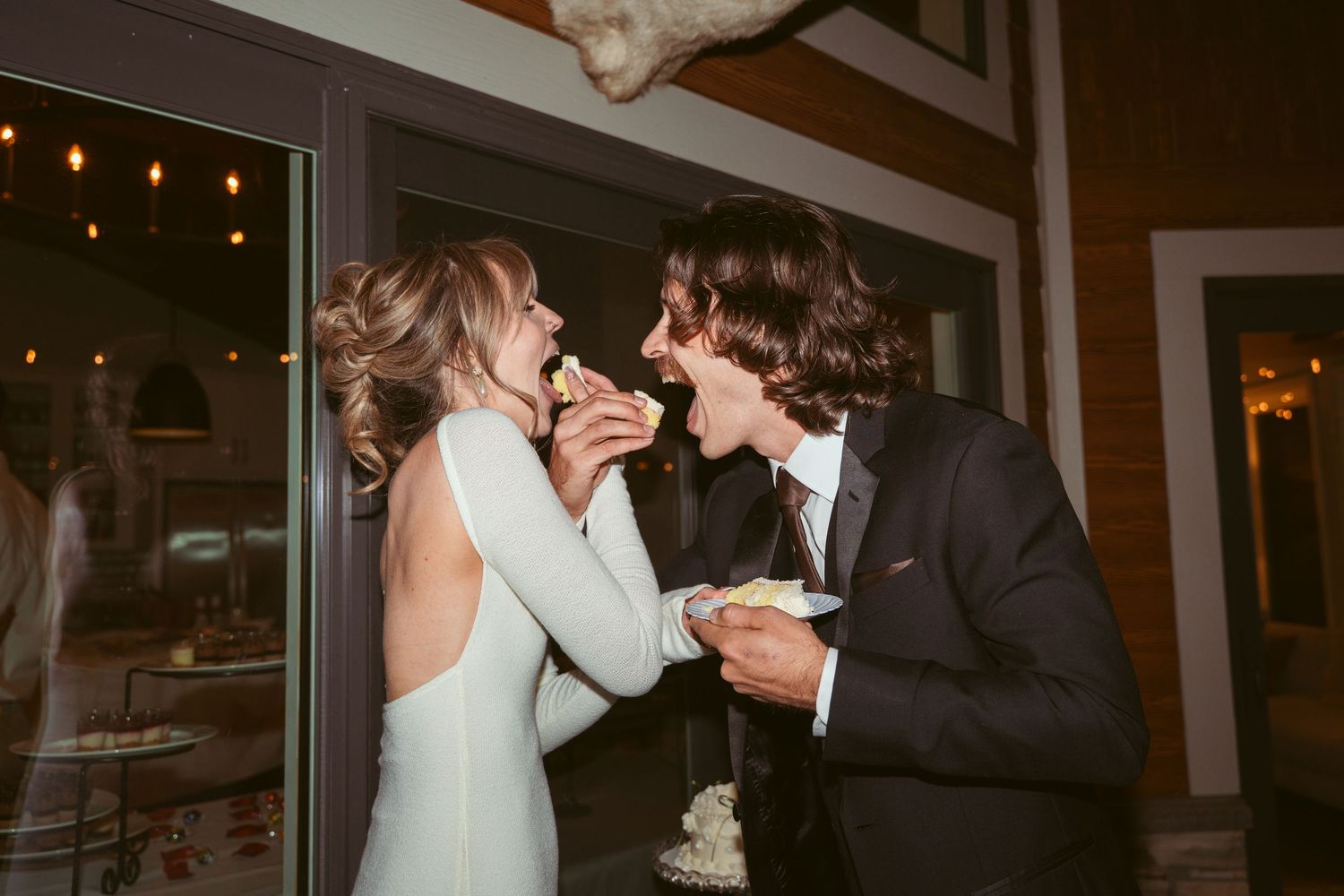Candid reception moment of newlyweds playfully sharing wedding cake in dim lighting.
