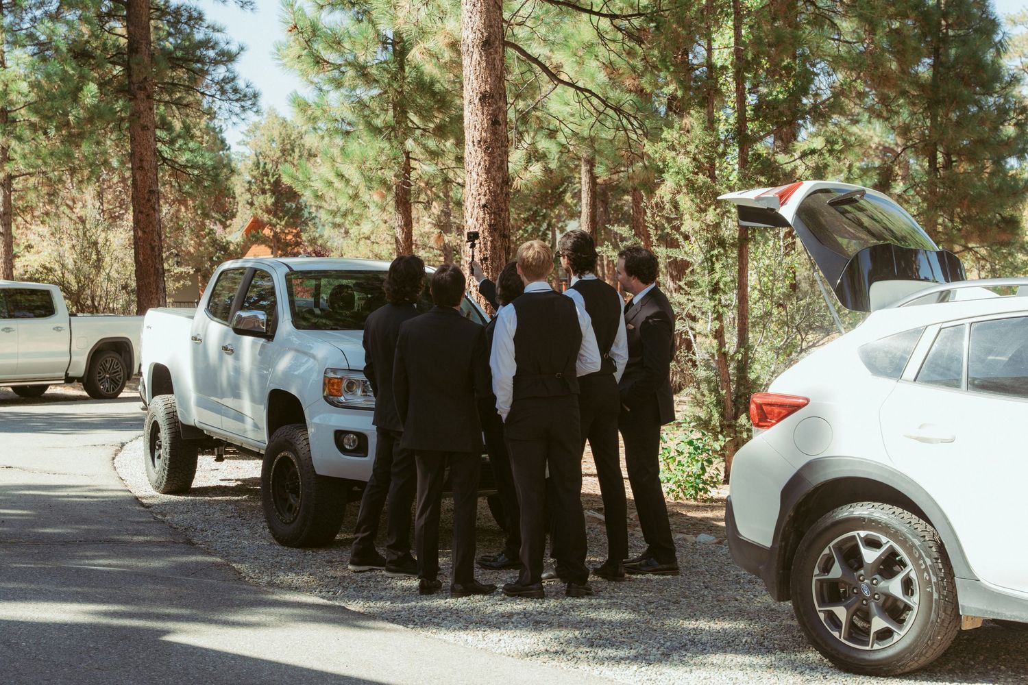 A group of people gather near parked cars on a tree-lined road during a wedding event.