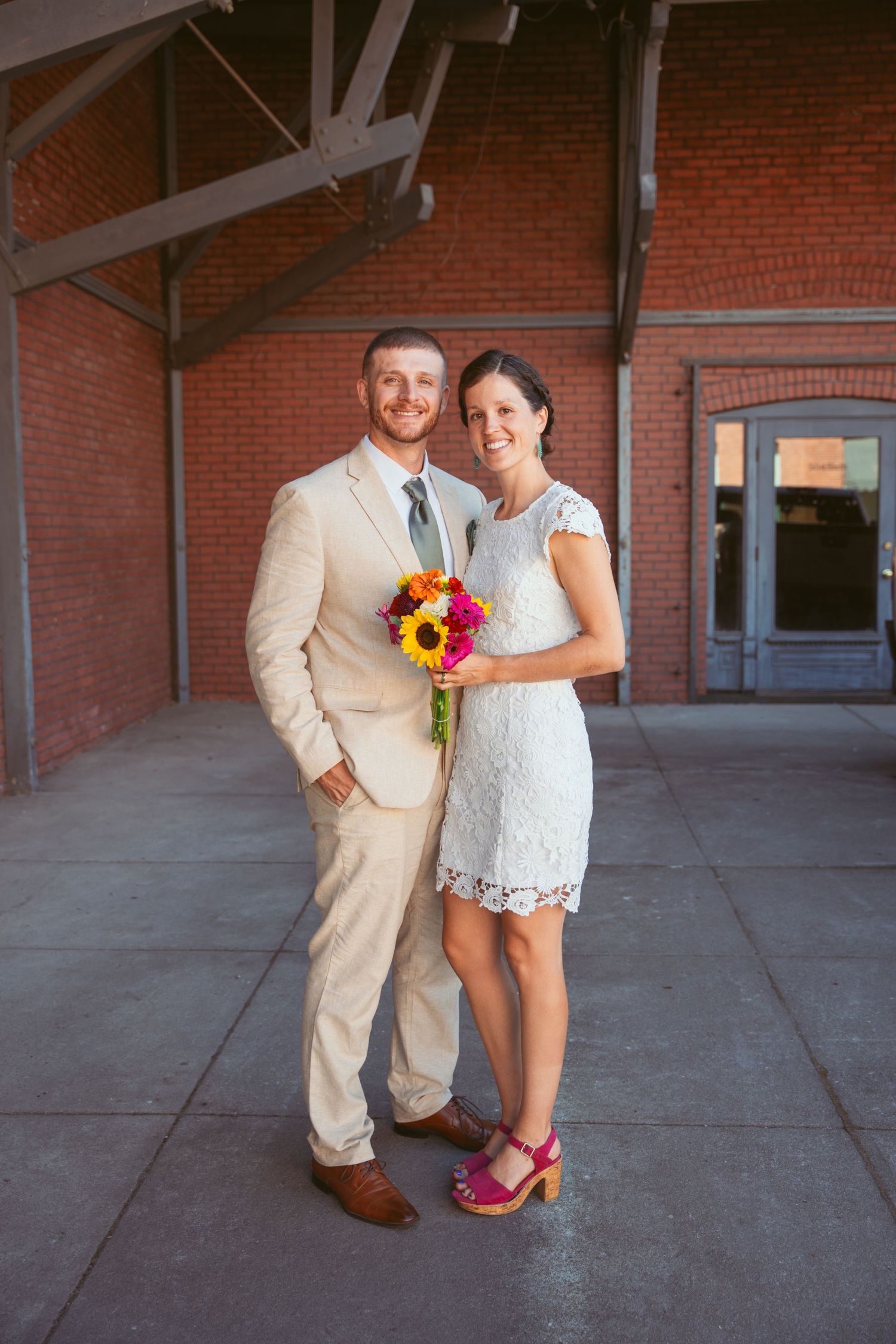 Romantic wedding couple poses against brick wall showing details of lace dress, tan suit, and colorful bouquet.