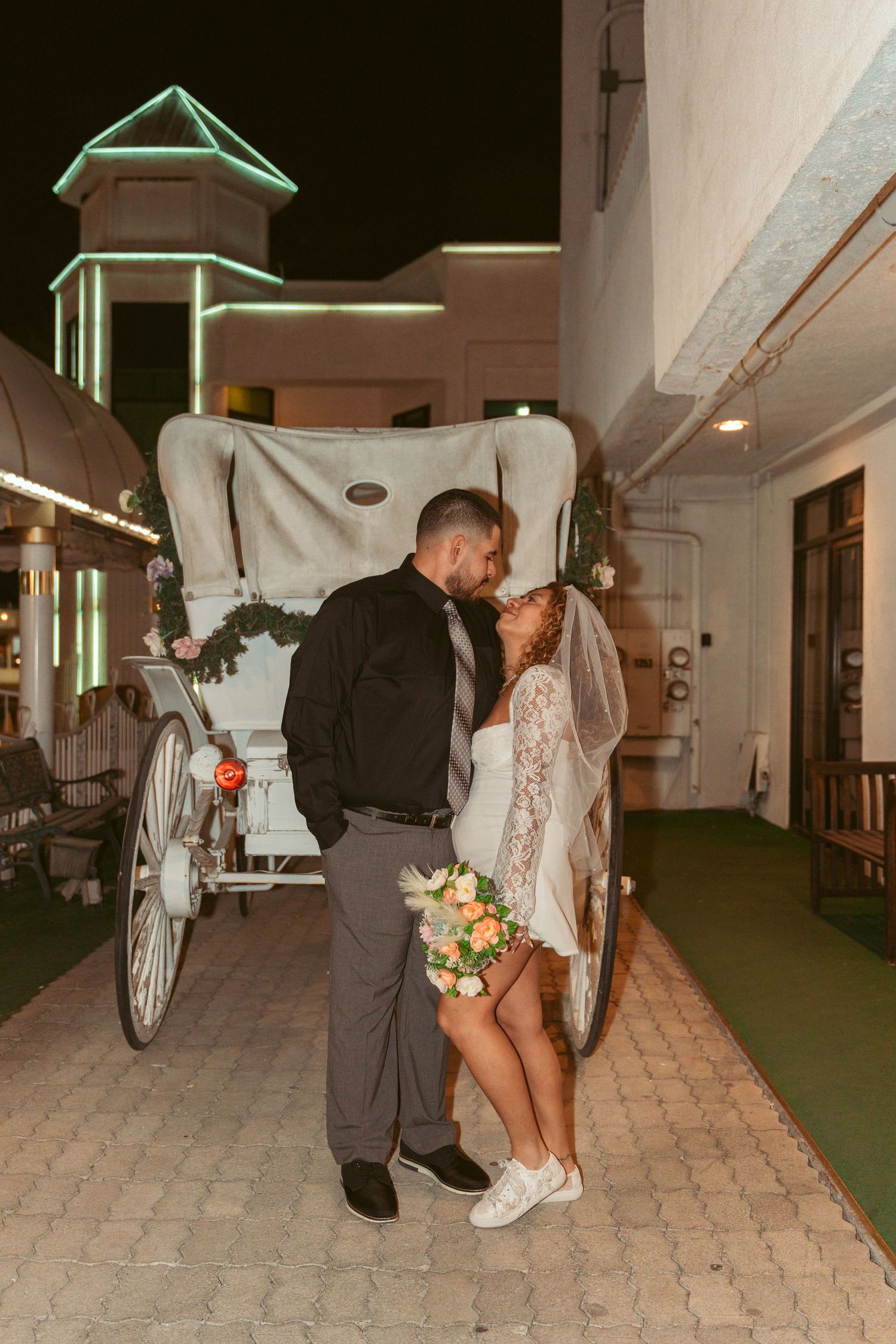 Couple posing near a decorative white carriage at a wedding venue with green accents.
