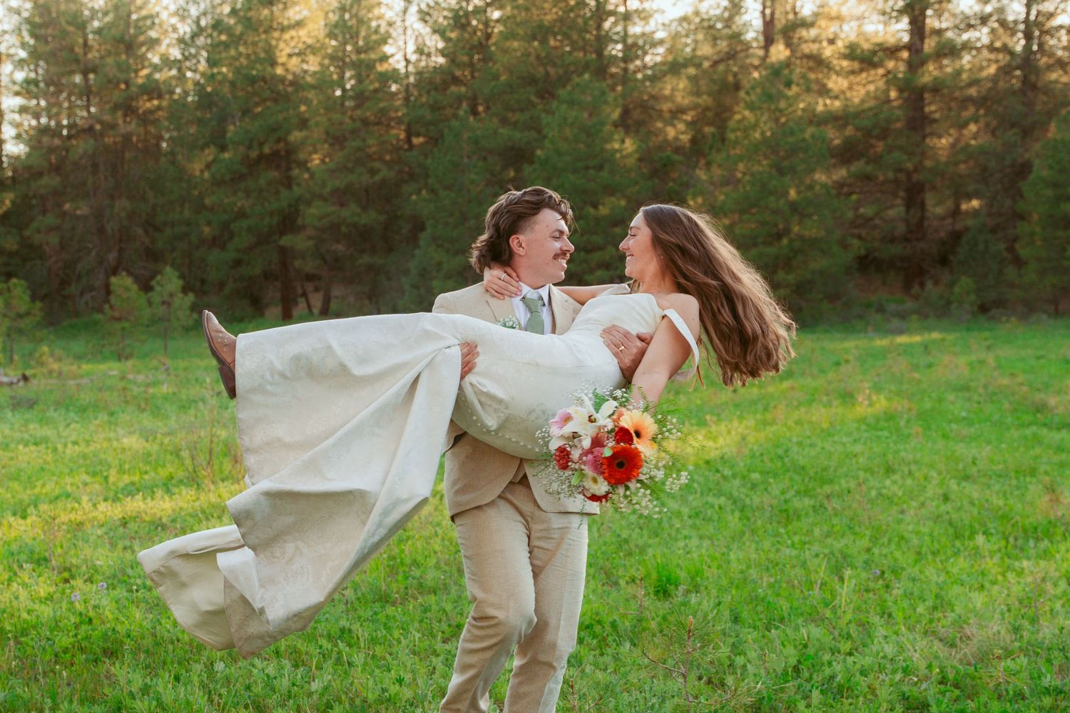 Playful couple dancing and twirling in a sunlit meadow with pine trees in the background.