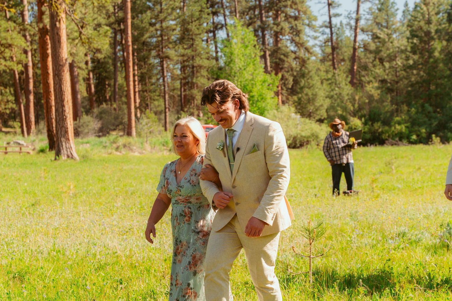 Wedding party walking and laughing in a grassy meadow with forest backdrop.