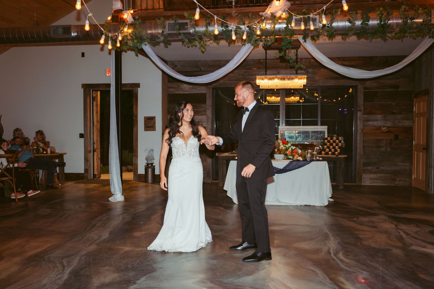 A couple shares their first dance at a rustic wedding reception with white draping and string lights overhead.