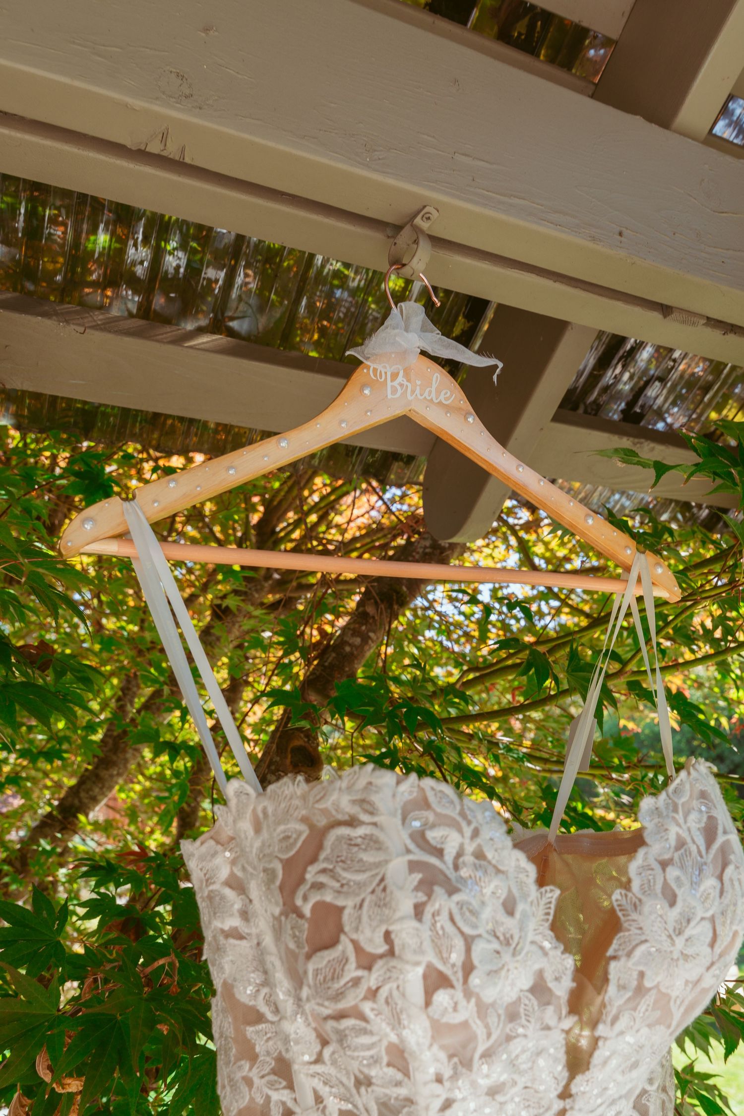 Close-up of lace wedding dress detail hanging on wooden hanger with sunlit foliage behind.