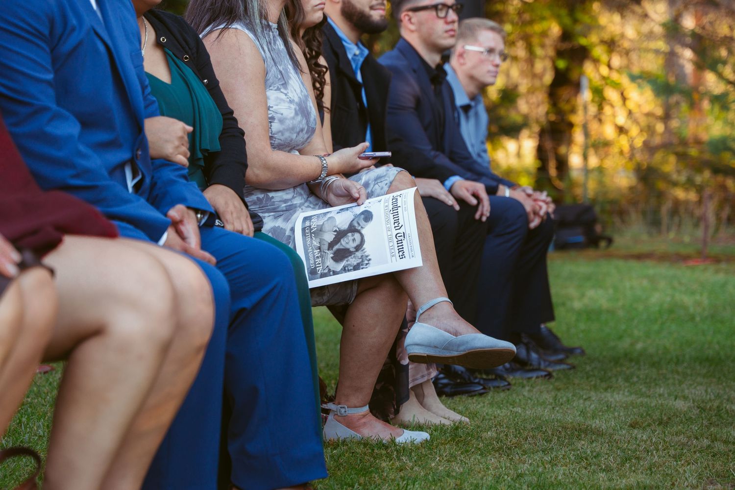 Wedding guests sit on benches outdoors holding programs during an autumn ceremony.
