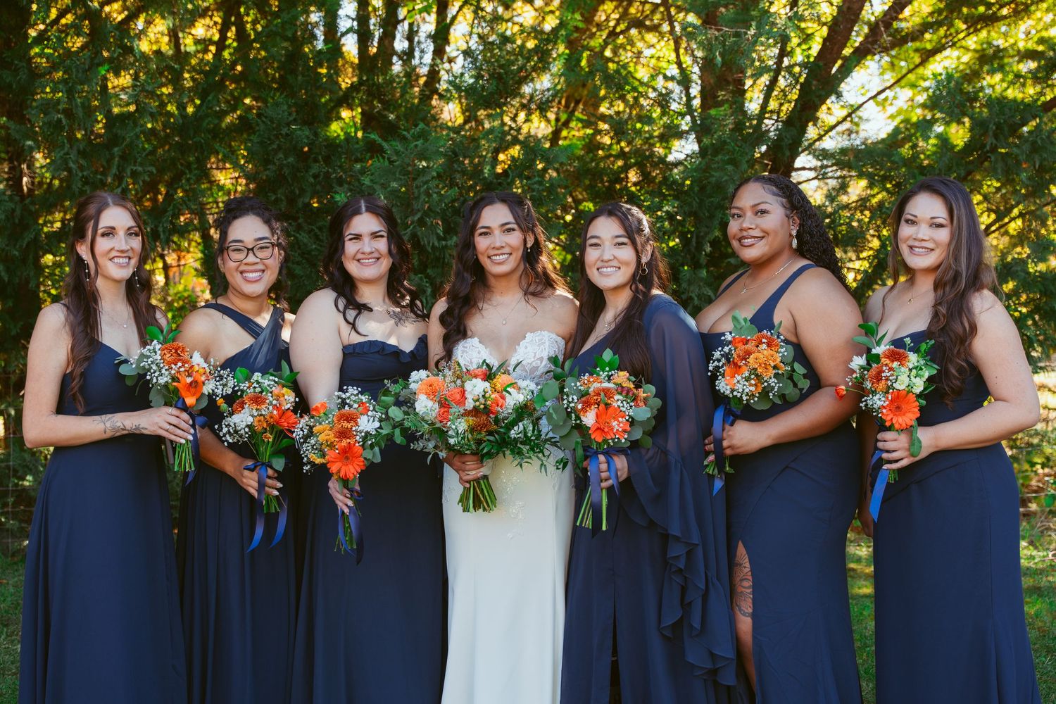 Wedding party wearing navy blue dresses holding vibrant orange and white bouquets in outdoor garden setting.