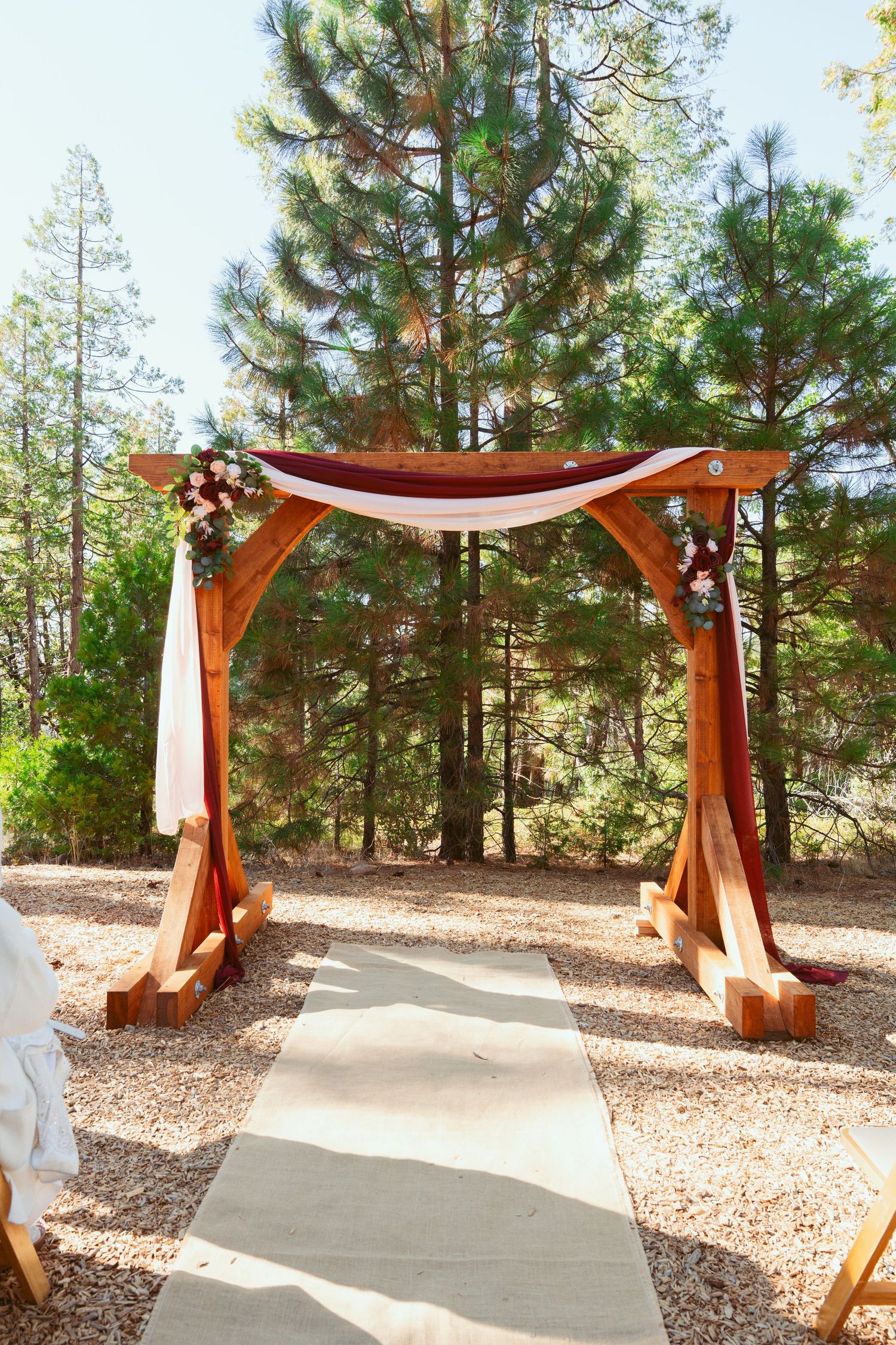 Wooden wedding arch with white fabric and floral decorations on forest ceremony path.
