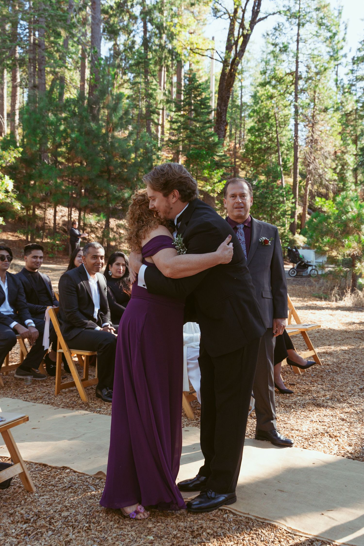 Wedding party members in formal attire interacting on a stone path surrounded by pine trees.