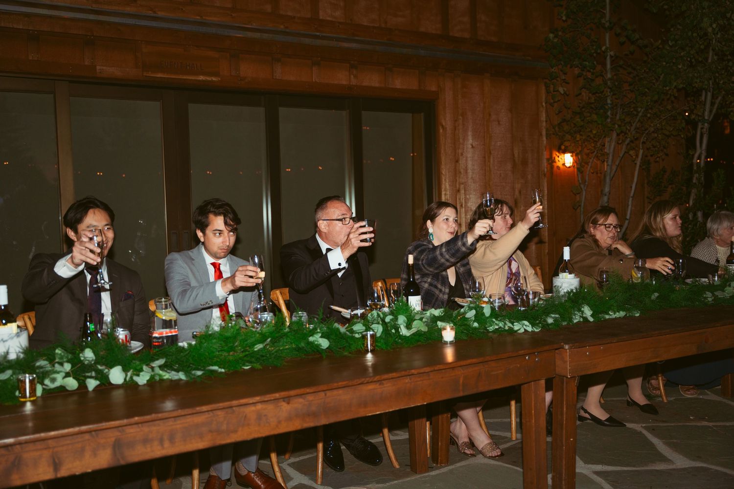 Wedding reception guests seated at long wooden tables in rustic outdoor venue.
