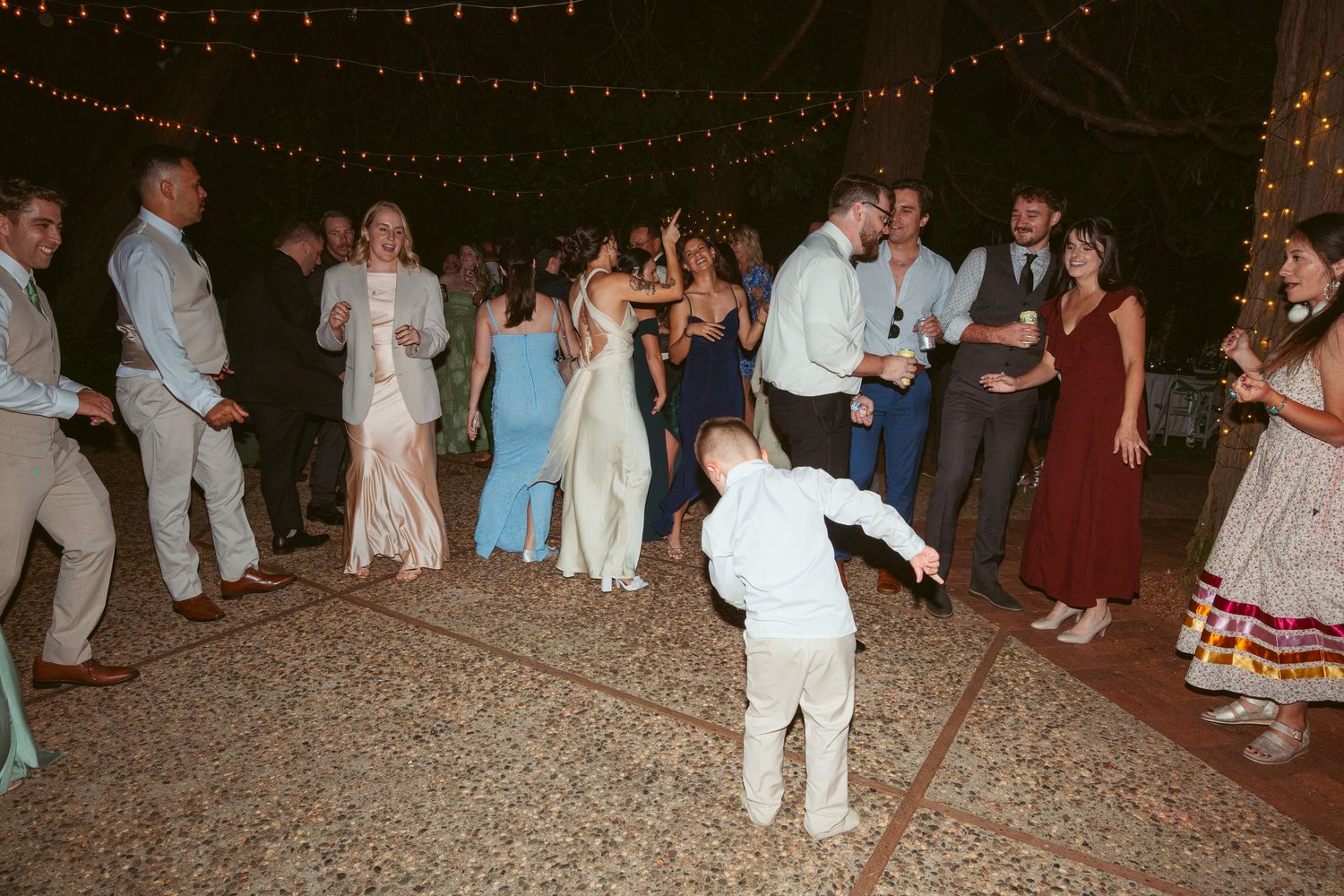 Wedding guests dance and celebrate at an outdoor reception with string lights overhead.