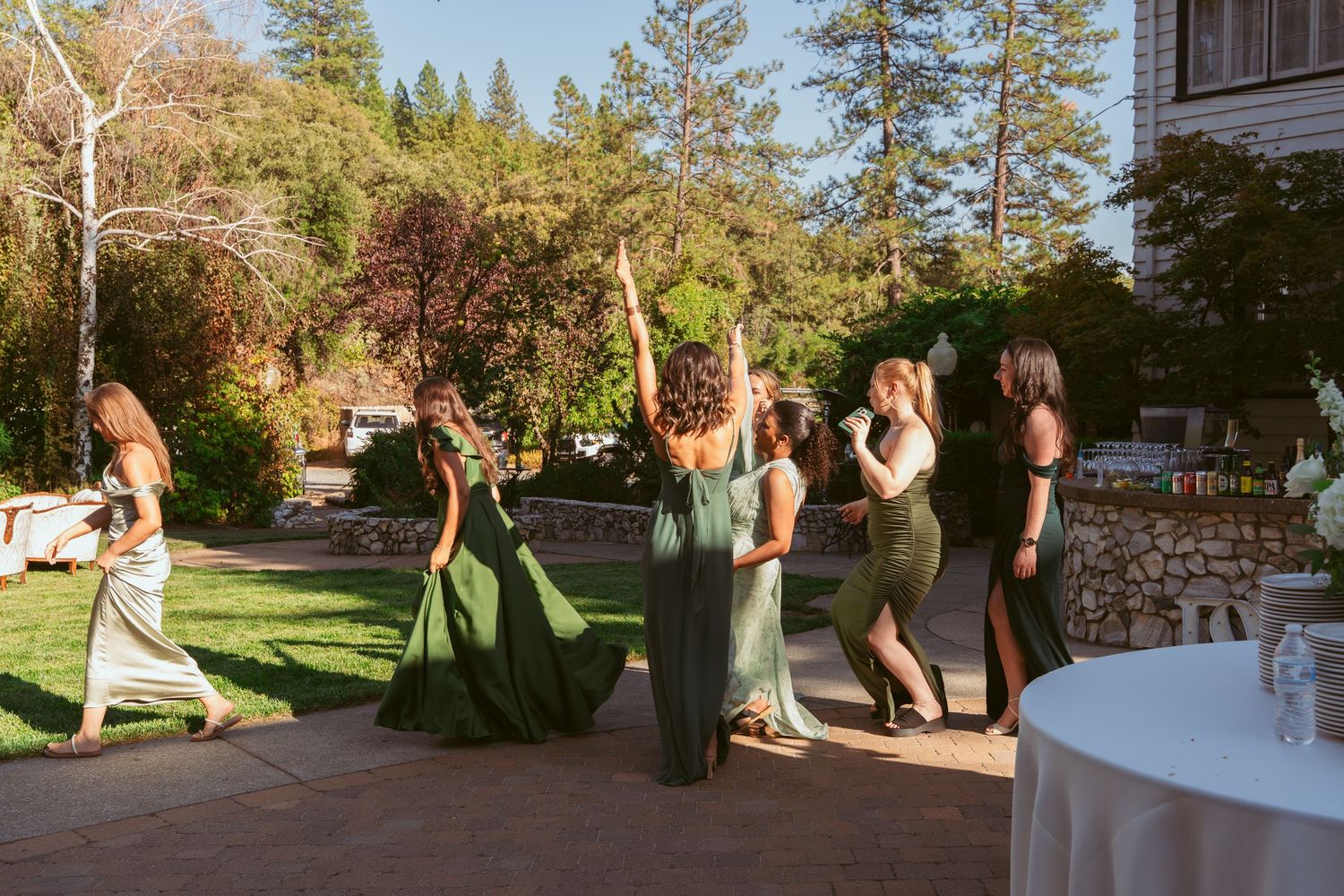 A group in formal attire gathers on a sunlit outdoor deck surrounded by trees during a special event.