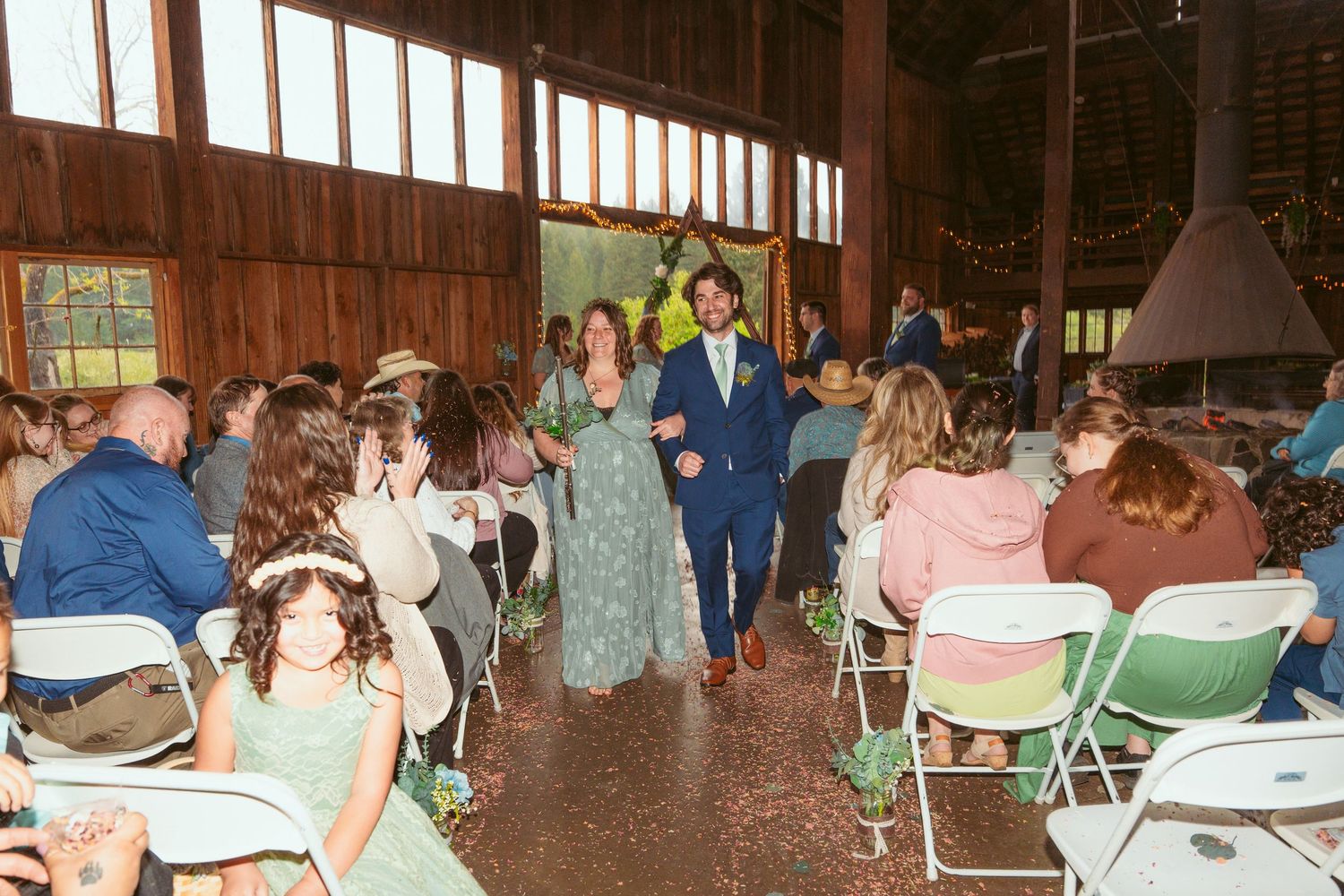 Wedding guests seated in folding chairs inside a rustic barn for a ceremony.