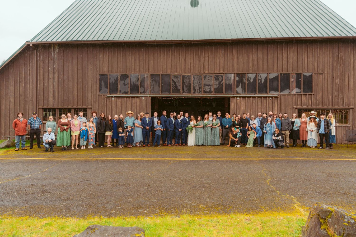 Large wedding group photo in front of rustic wooden barn with green hills in background.