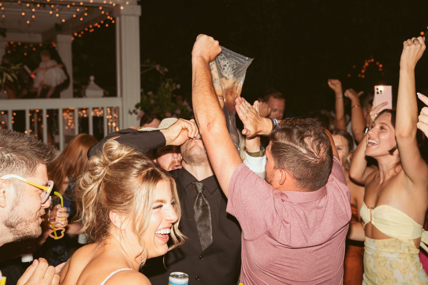 Energetic crowd dancing under string lights at wedding reception party.