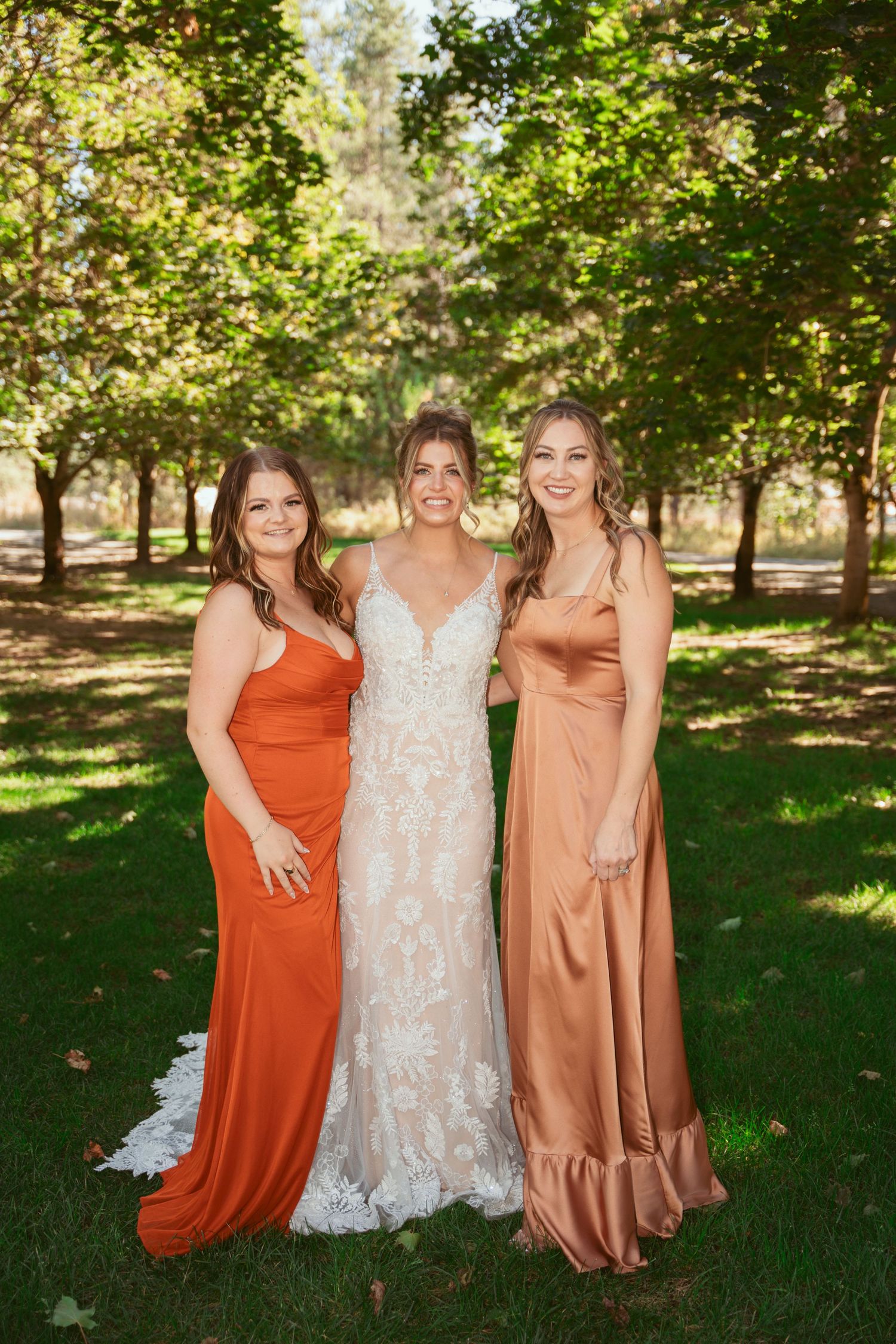 Bridal party in orange and copper dresses pose together on green lawn.