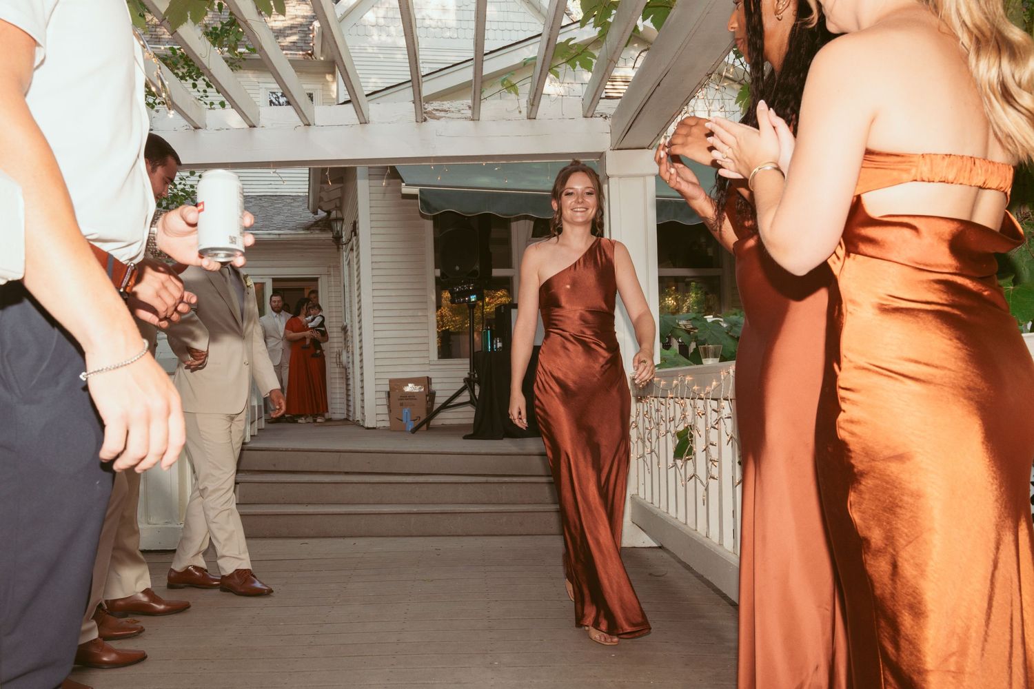 Wedding guests dancing in formal attire with orange bridesmaid dresses visible on a covered outdoor patio.
