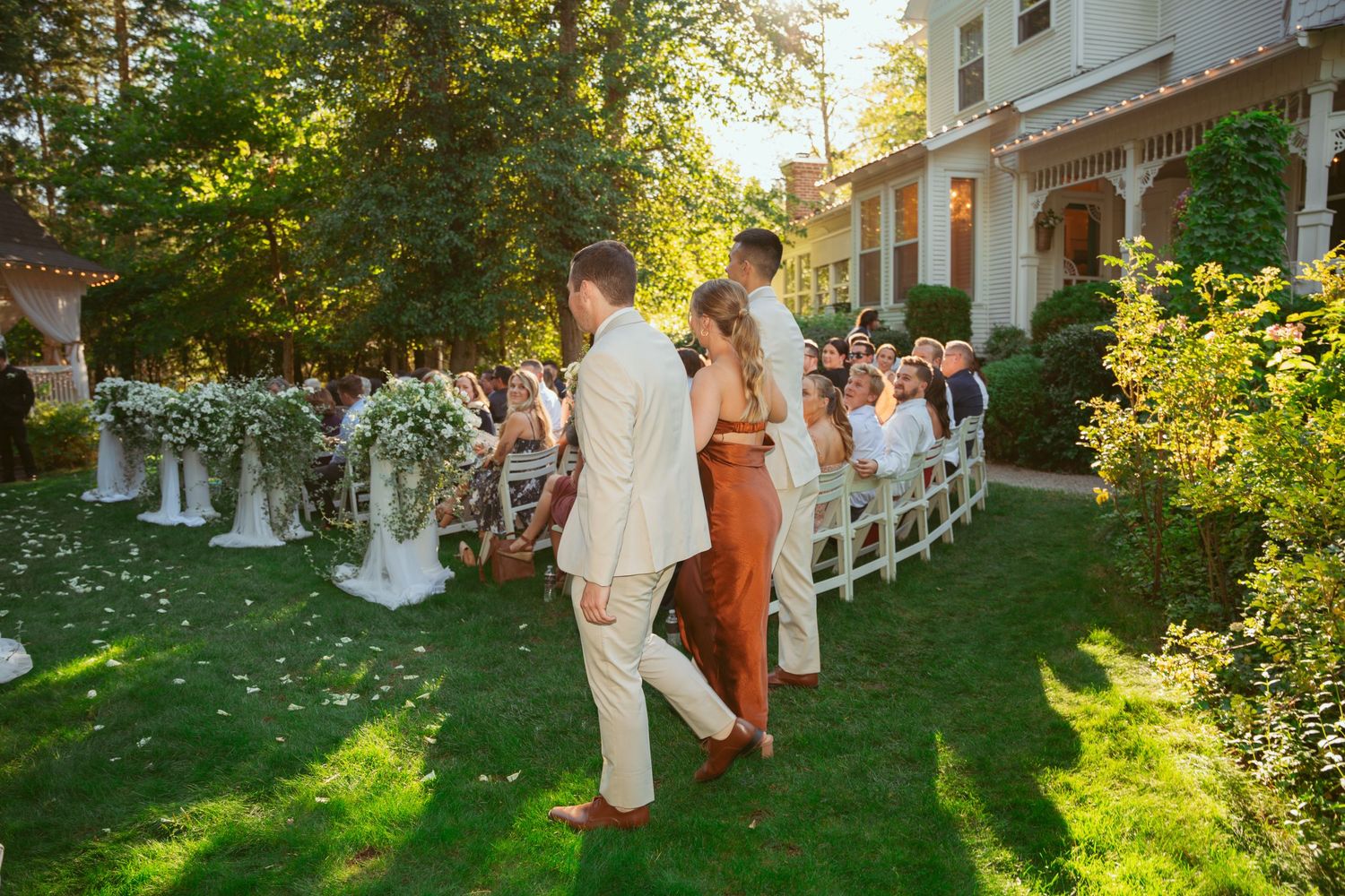 Wedding guests seated outdoors on white chairs as couple walks down garden aisle at sunset.