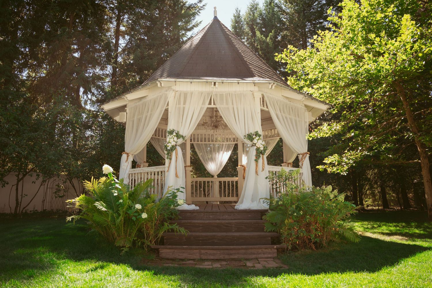 White outdoor wedding gazebo setup surrounded by lush green trees and landscaping.