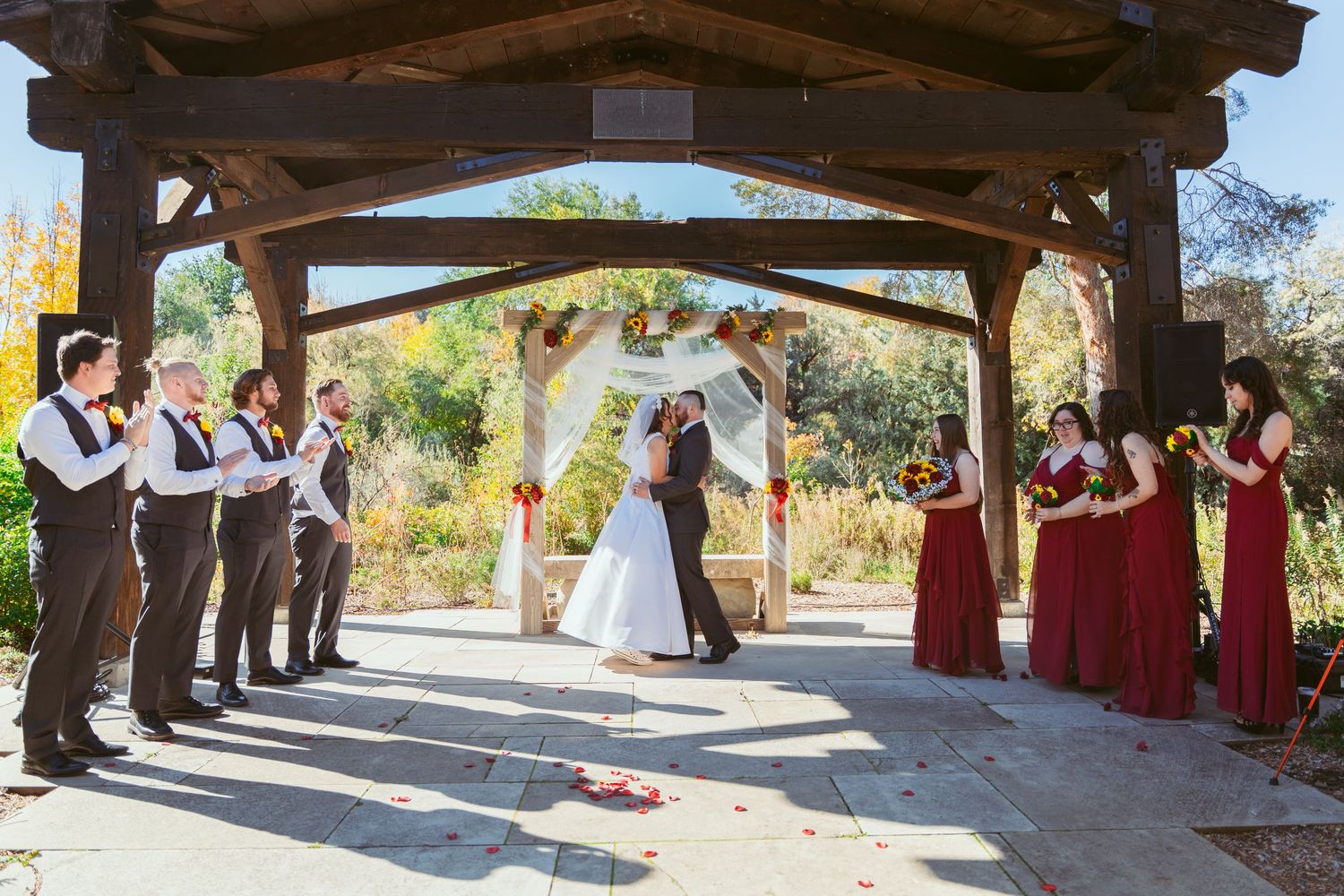 Panoramic view of a wedding ceremony with attendants in burgundy dresses under a stone pavilion.