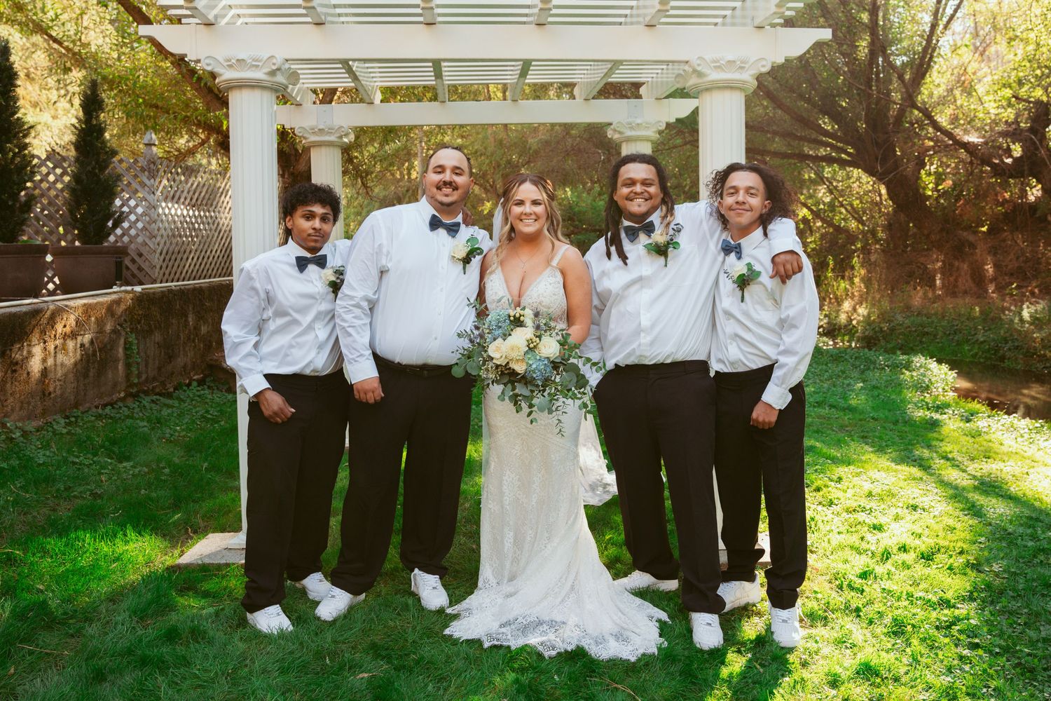 Wedding party poses under a white pergola in a garden setting with groomsmen in light shirts.