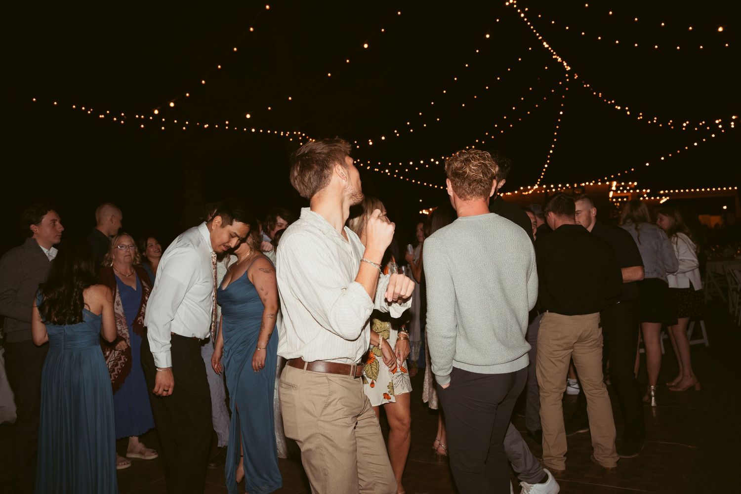 Wedding guests mingle and dance under strings of outdoor reception lighting.