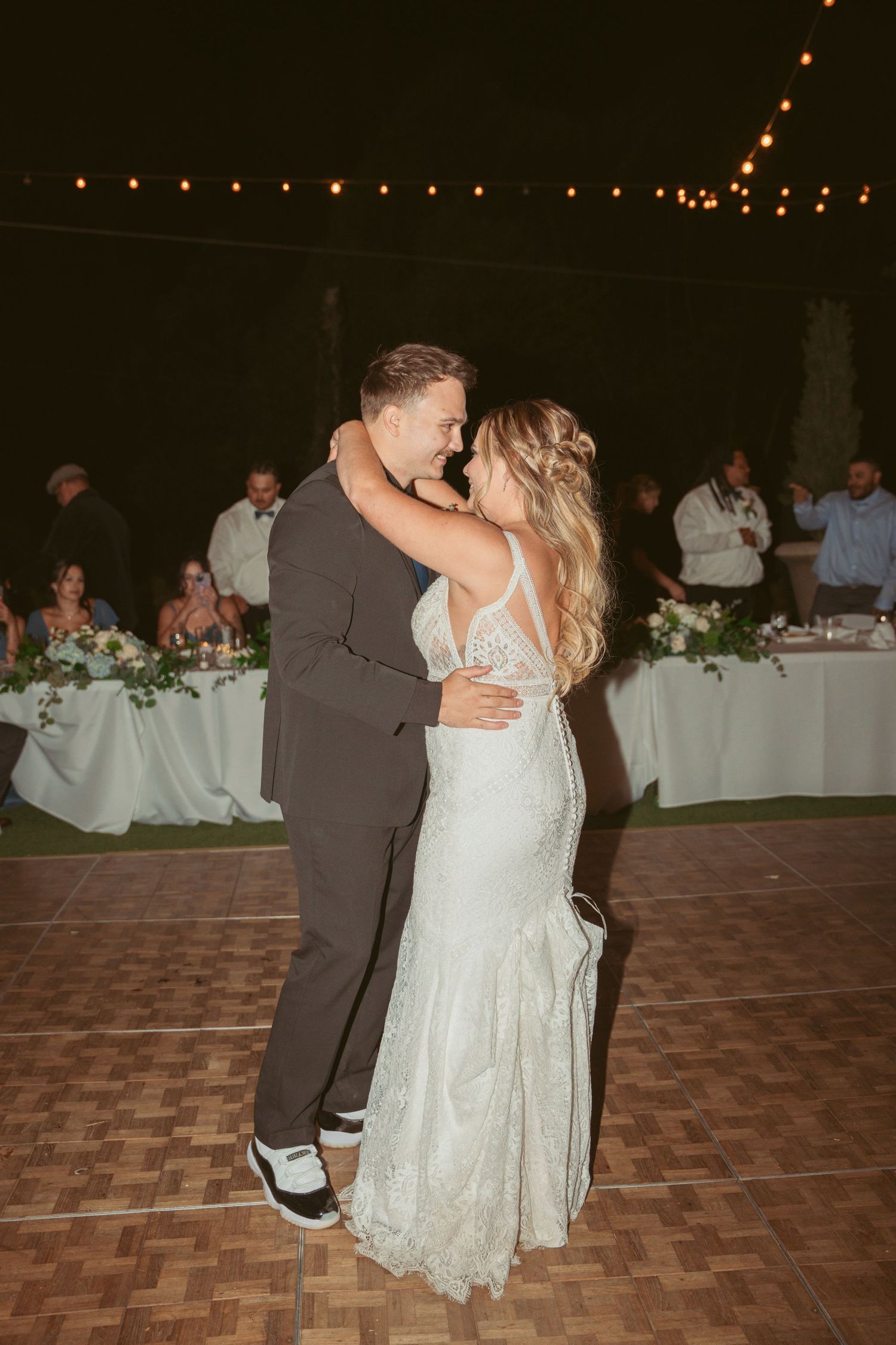 A couple shares an intimate dance on a wooden floor at an outdoor reception with string lights overhead.