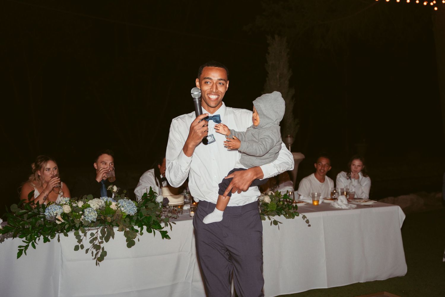 Someone in formal attire holding a small child during what appears to be a wedding reception.